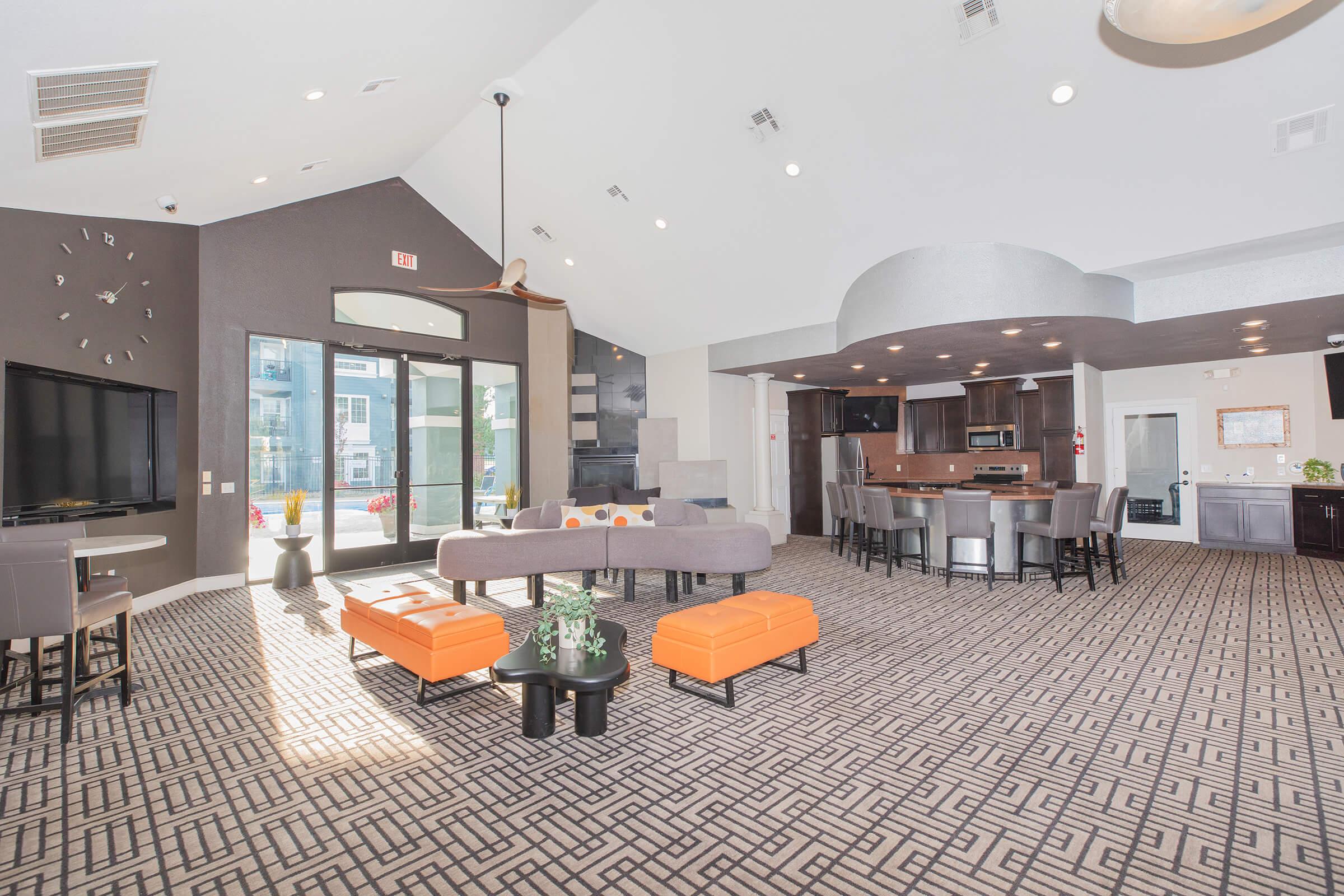 A modern lounge area featuring stylish furniture, including gray sofas and orange accent tables. Large windows provide natural light, showcasing a spacious, open design with a patterned carpet. A bar area is visible in the background, complemented by contemporary decor and lighting fixtures.