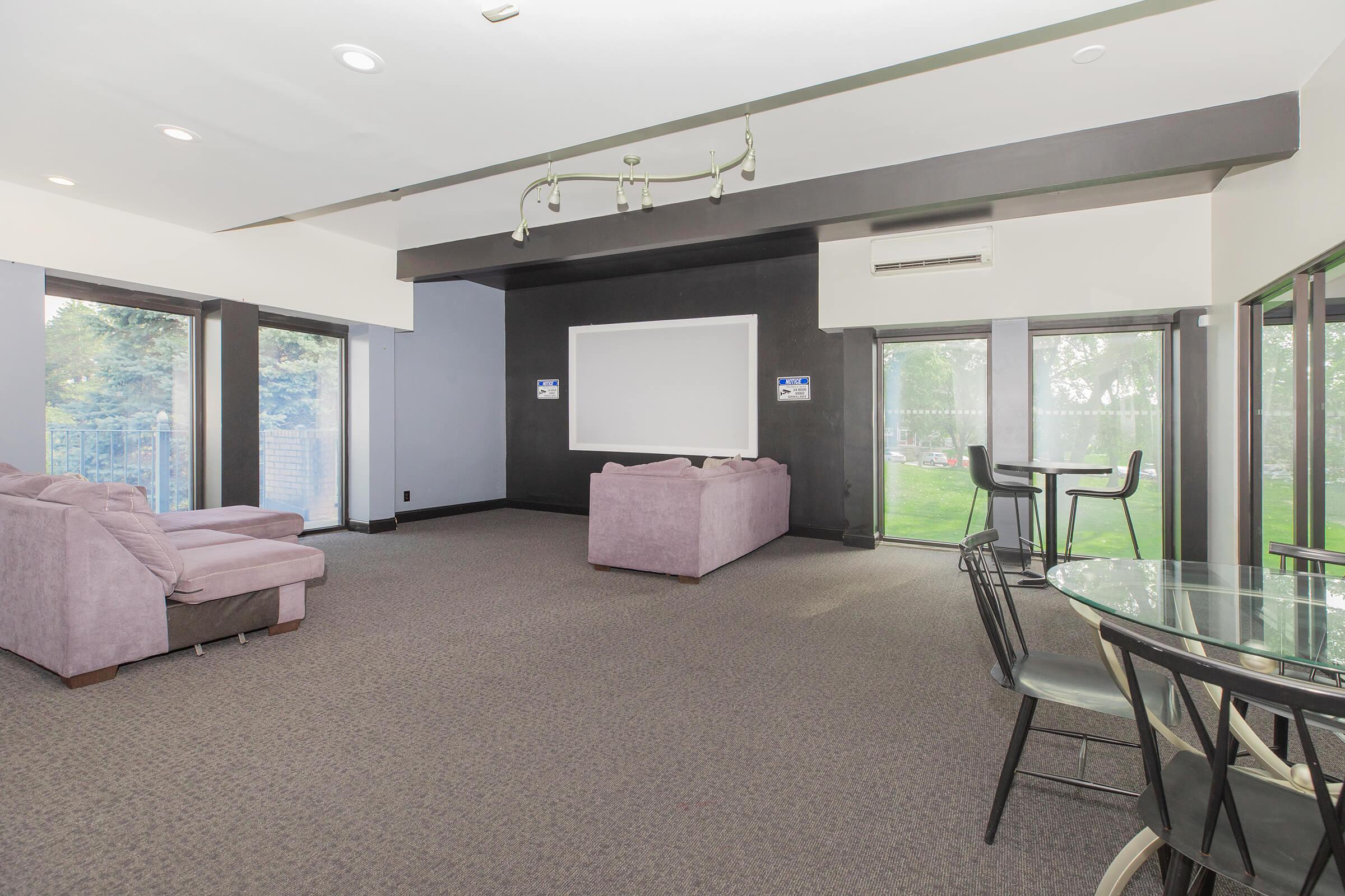 A spacious interior room featuring a seating area with purple sofas, a central blank whiteboard on a black wall, and large windows that open to a green outdoor space. There are also a dining table with black chairs and a carpeted floor, creating a comfortable and inviting atmosphere.