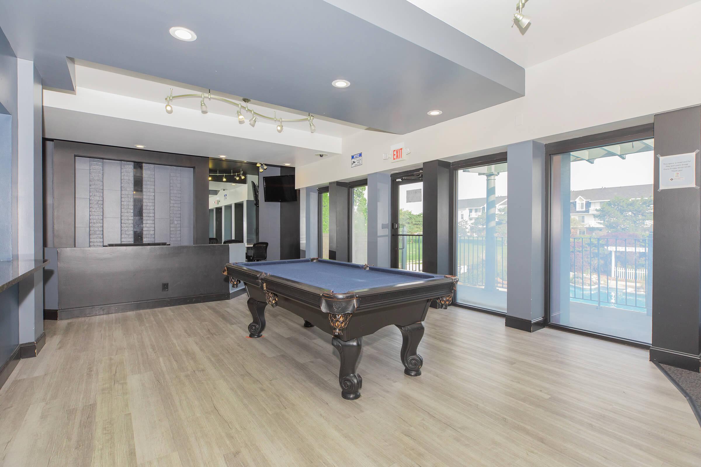 A modern recreational room featuring a black pool table with a blue felt surface. The space has large windows providing natural light, wooden flooring, and a stylish decor, creating a welcoming atmosphere for relaxation and socializing. A television is mounted on the wall, adding to the entertainment options.