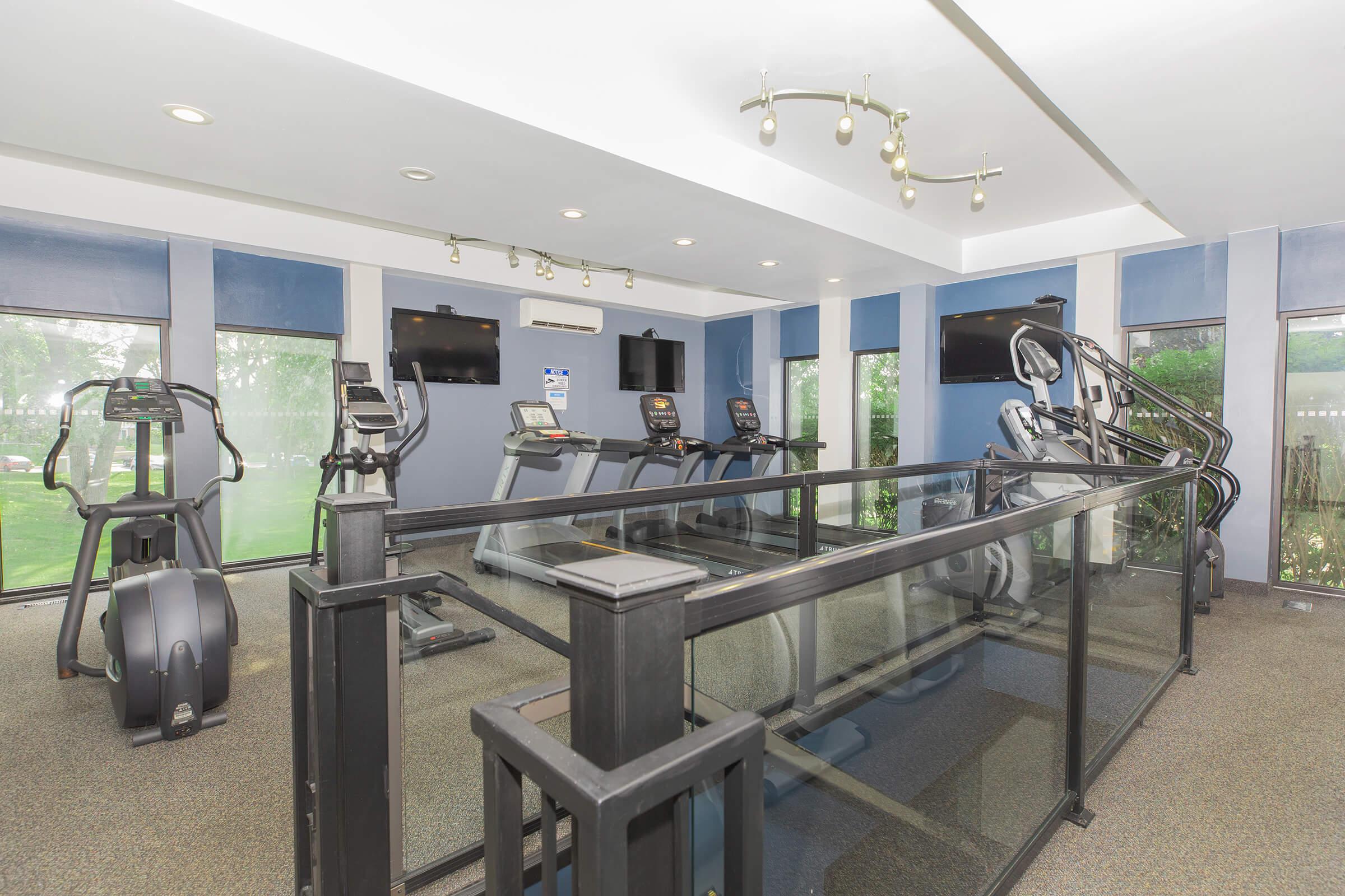 A modern gym interior featuring multiple exercise machines, including elliptical trainers and treadmills. The space is well-lit with large windows providing views of greenery outside. The walls are painted in a calming blue color, and there are two television screens mounted on the walls for entertainment during workouts.