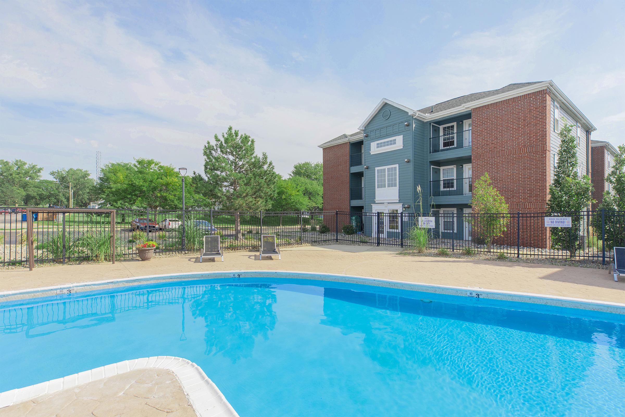 A clear blue swimming pool in the foreground, surrounded by a sandy area and lounge chairs. In the background, a three-story brick and blue apartment building is visible, along with green trees and landscaping. The sky is bright and partly cloudy, creating a pleasant outdoor setting.