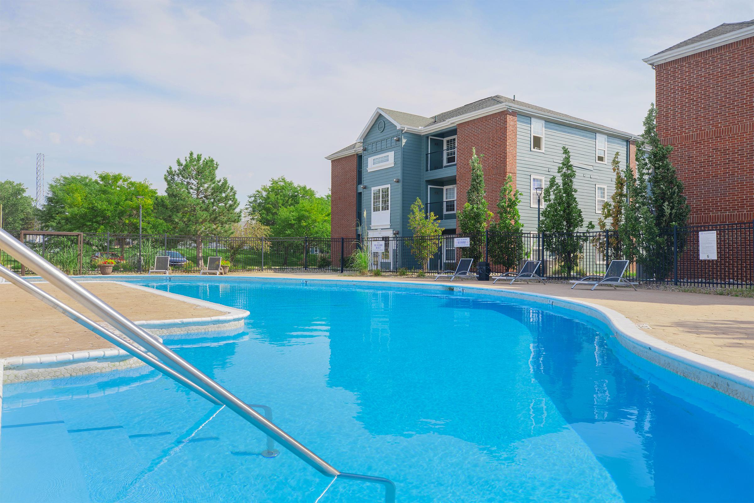 A swimming pool surrounded by several trees and lounge chairs, with a brick apartment building in the background. The pool features a gentle curve and a railing leading into the water. The scene is bright and sunny, creating a relaxing atmosphere.