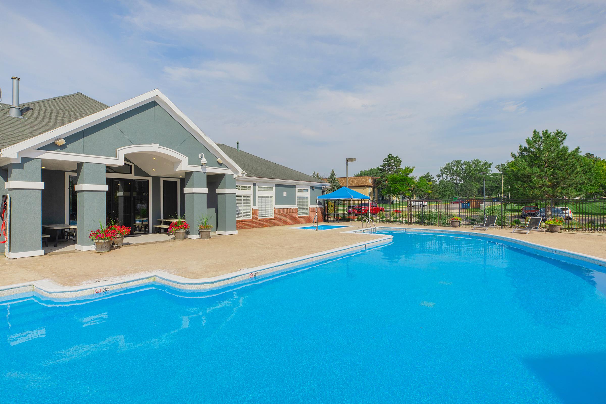 A sparkling blue swimming pool surrounded by a relaxing deck, with potted plants and a nearby shaded area. An inviting building with large windows overlooks the pool area. Green trees and a fence are visible in the background, creating a serene outdoor atmosphere.