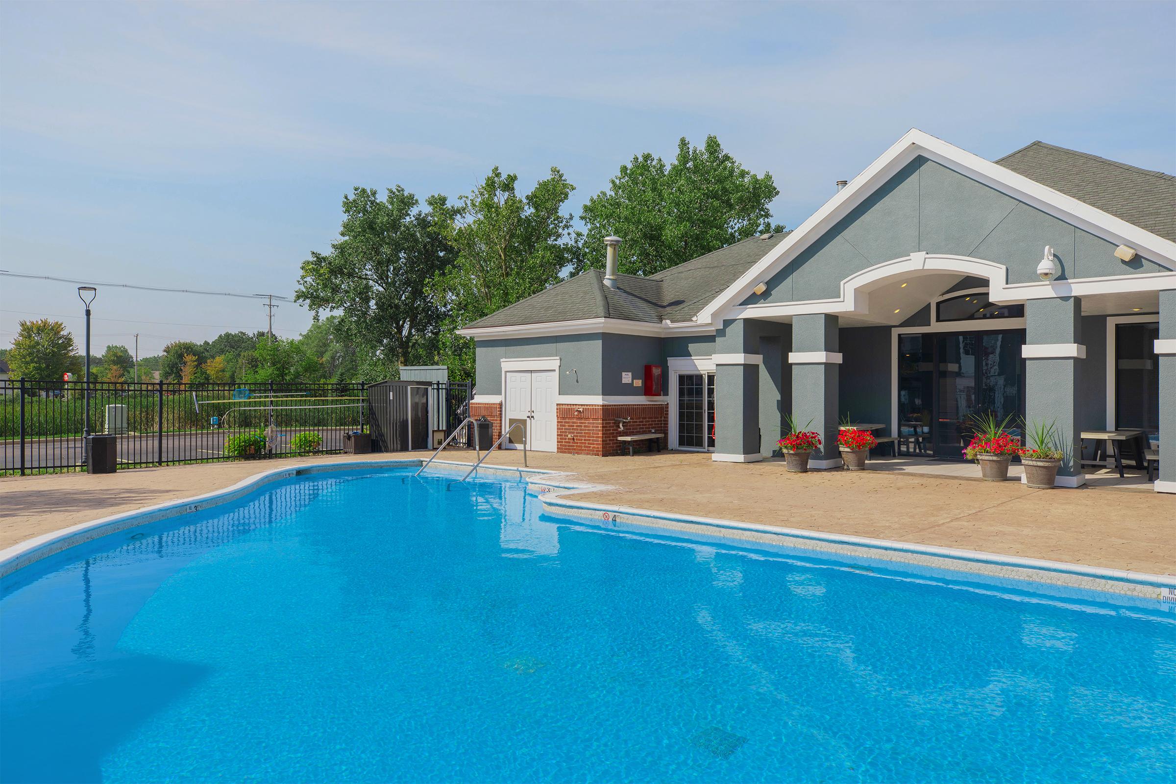 A clear blue swimming pool with a well-maintained patio area. To the right, there's a modern building with large windows, featuring a light gray exterior and potted plants. Trees are visible in the background, and the sky is partly cloudy, creating a serene outdoor atmosphere.