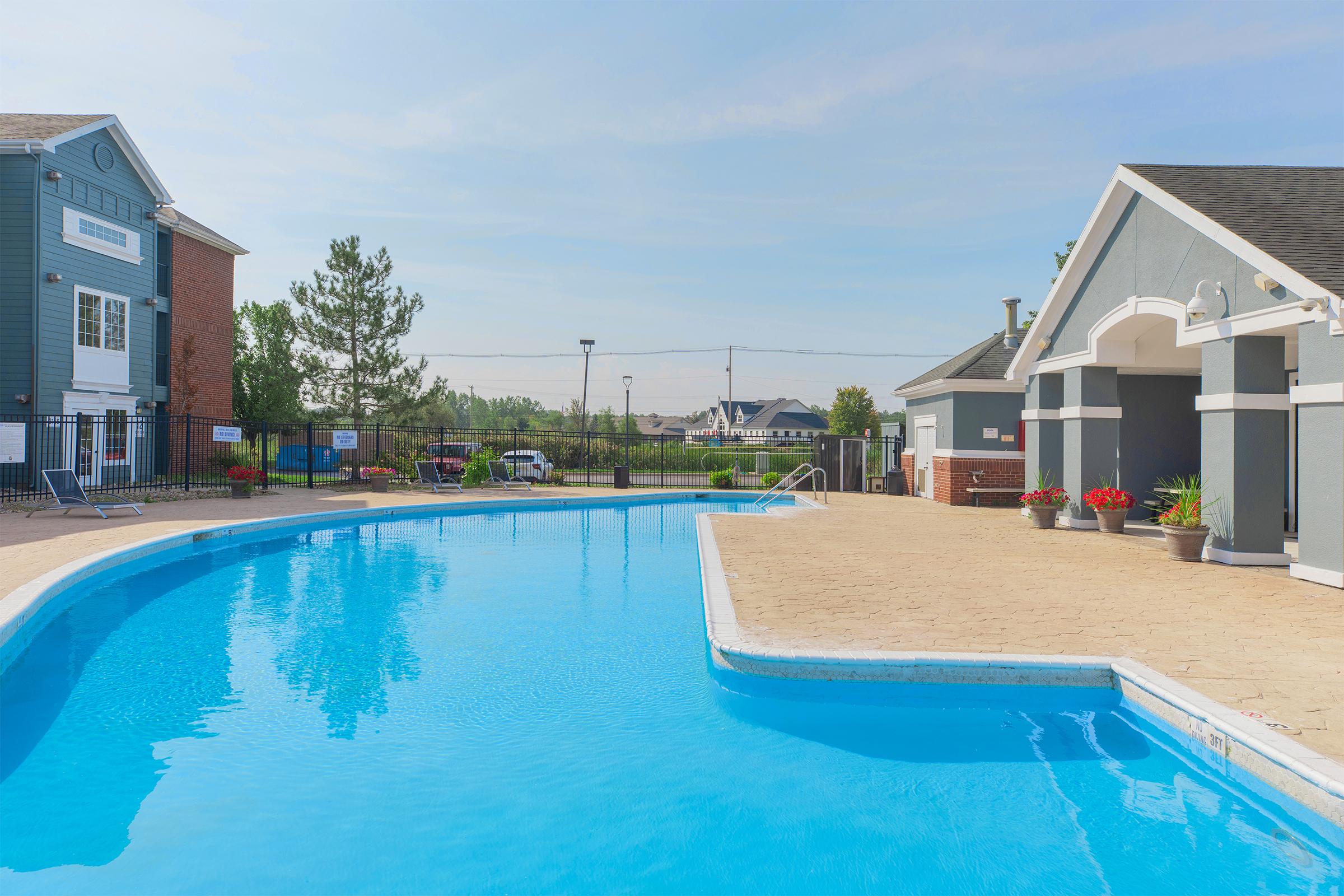 A sparkling blue swimming pool surrounded by a patio area, with lounge chairs on one side. Nearby, there are two buildings with gray and brick exteriors. The scene is bright and sunny, with trees and landscaping in the background, creating a relaxing outdoor environment.
