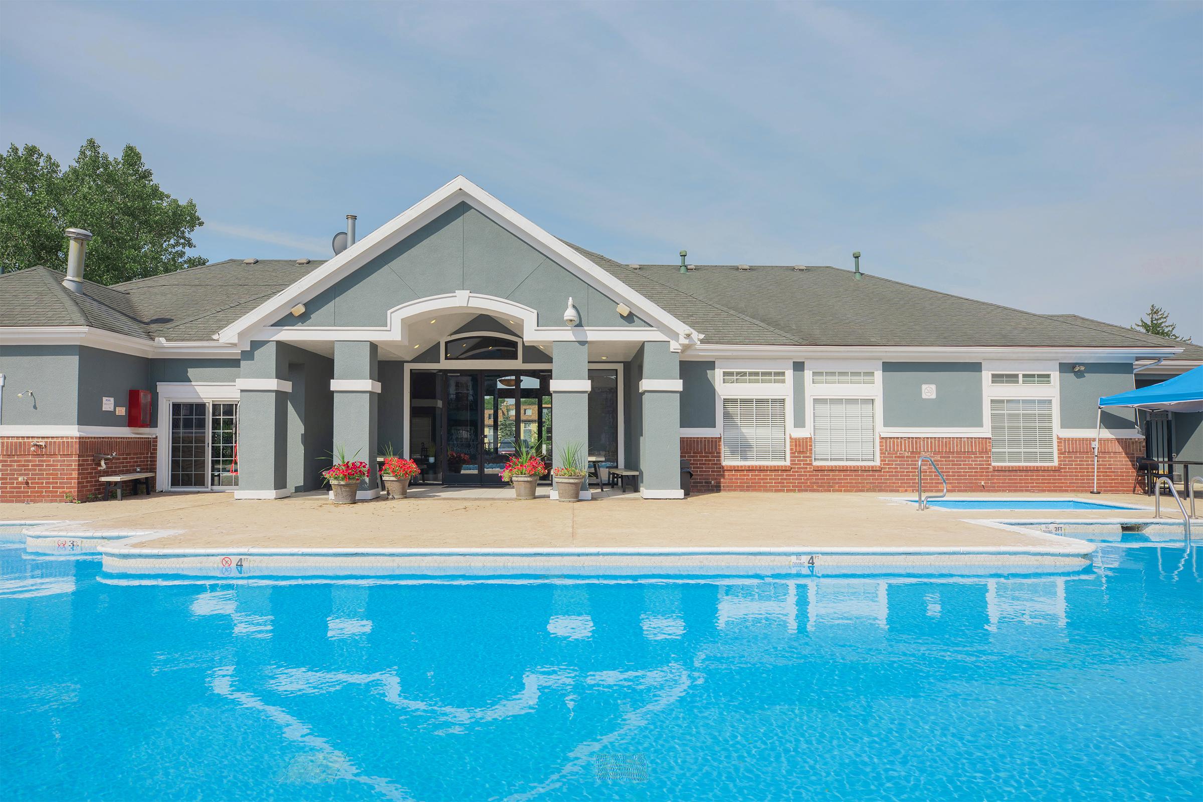 A sunny outdoor pool area featuring clear blue water, surrounded by a sandy area and planters with blooming flowers. In the background, there is a large, modern building with large windows and a neatly landscaped exterior. The sky is partly cloudy, creating a bright atmosphere.
