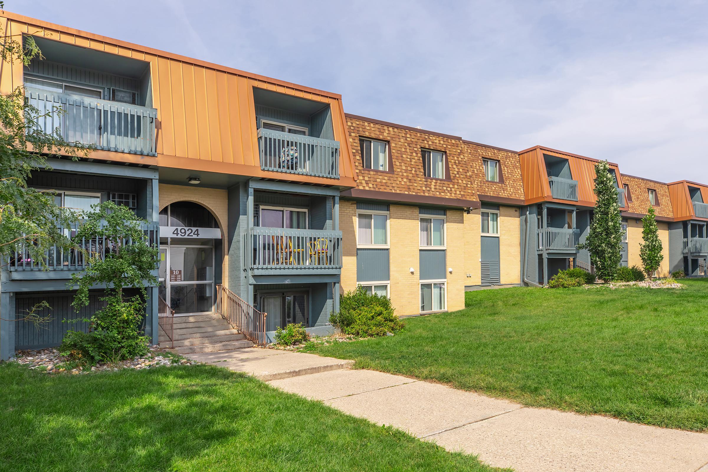 Row of multi-story apartment buildings with a green lawn in front. Each building features balconies with railings. The exterior has a mix of colors, including beige and orange roofing. A concrete pathway leads to the entrance of the leftmost building, and trees are visible on either side.