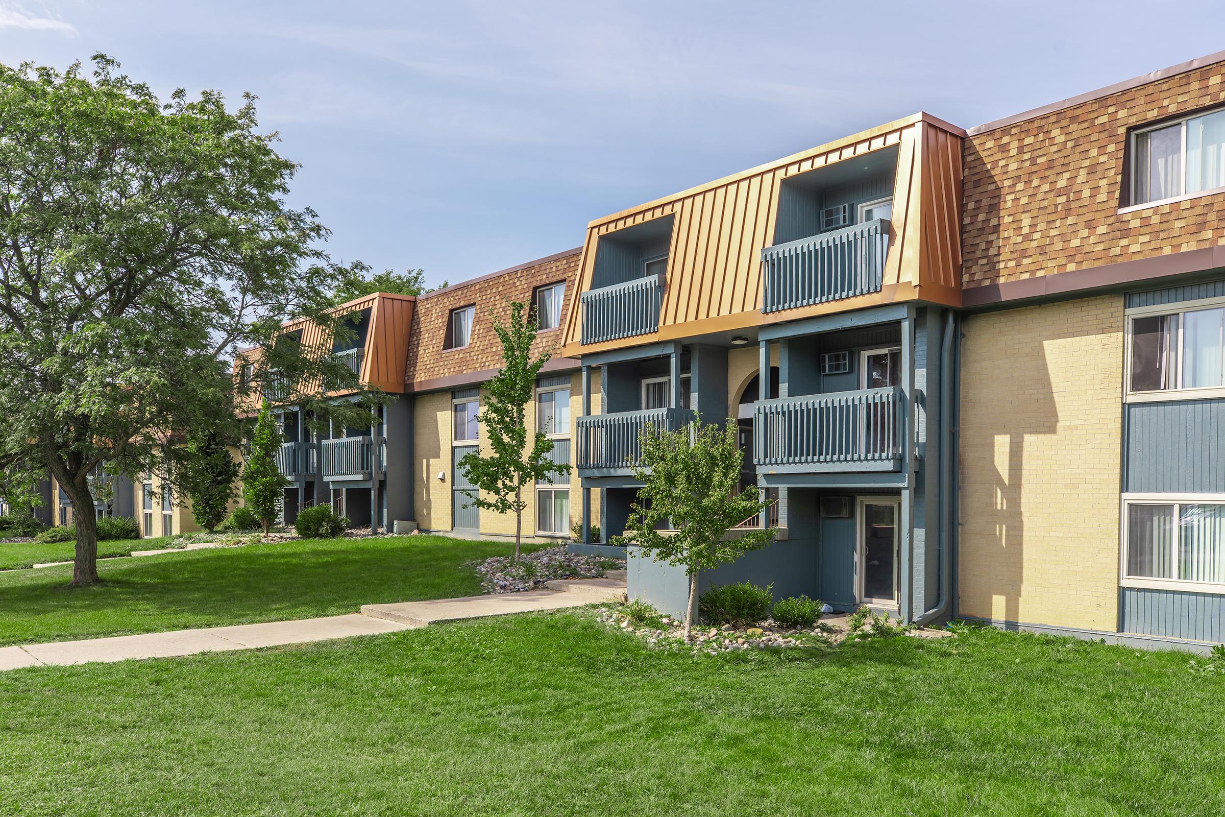 A row of two-story apartment buildings with balconies and a mix of brick and siding exteriors. Lush green grass and well-maintained bushes surround the buildings, and a tree is visible to the left. The sky is clear with a few clouds. The setting appears peaceful and residential.
