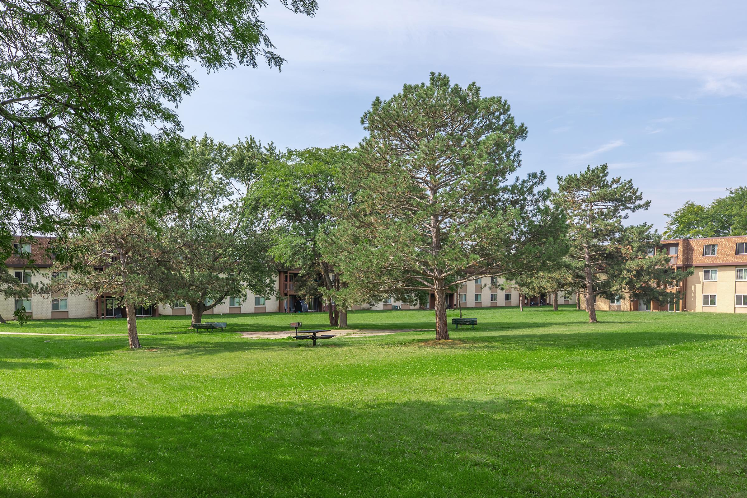 A spacious green lawn surrounded by trees, with a park bench positioned near a small gathering area. In the background, there are multiple buildings with balconies, set against a clear blue sky. The scene conveys a peaceful and inviting outdoor environment.