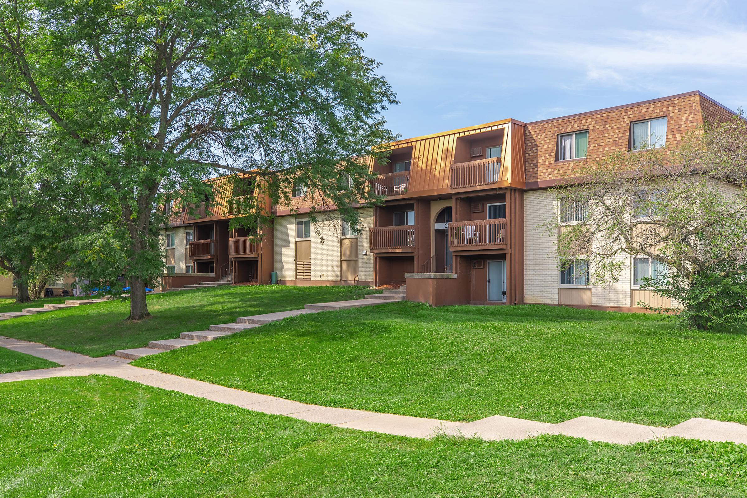 A view of a multi-unit residential building surrounded by green grass and trees. The building features multiple balconies, light-colored brick walls, and a brown roof. A paved walkway leads through the lawn, connecting the entrances to the apartments. The sky is clear with a few clouds.