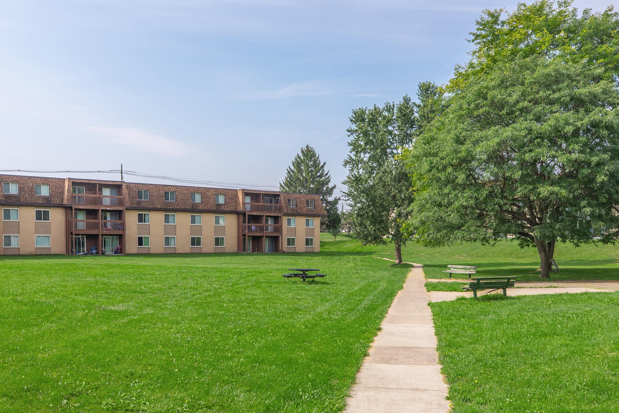A grassy area with two benches along a walkway leading to a row of low-rise apartment buildings. The buildings have balconies, and trees are visible nearby. The sky is clear with a few clouds, creating a bright, inviting environment.