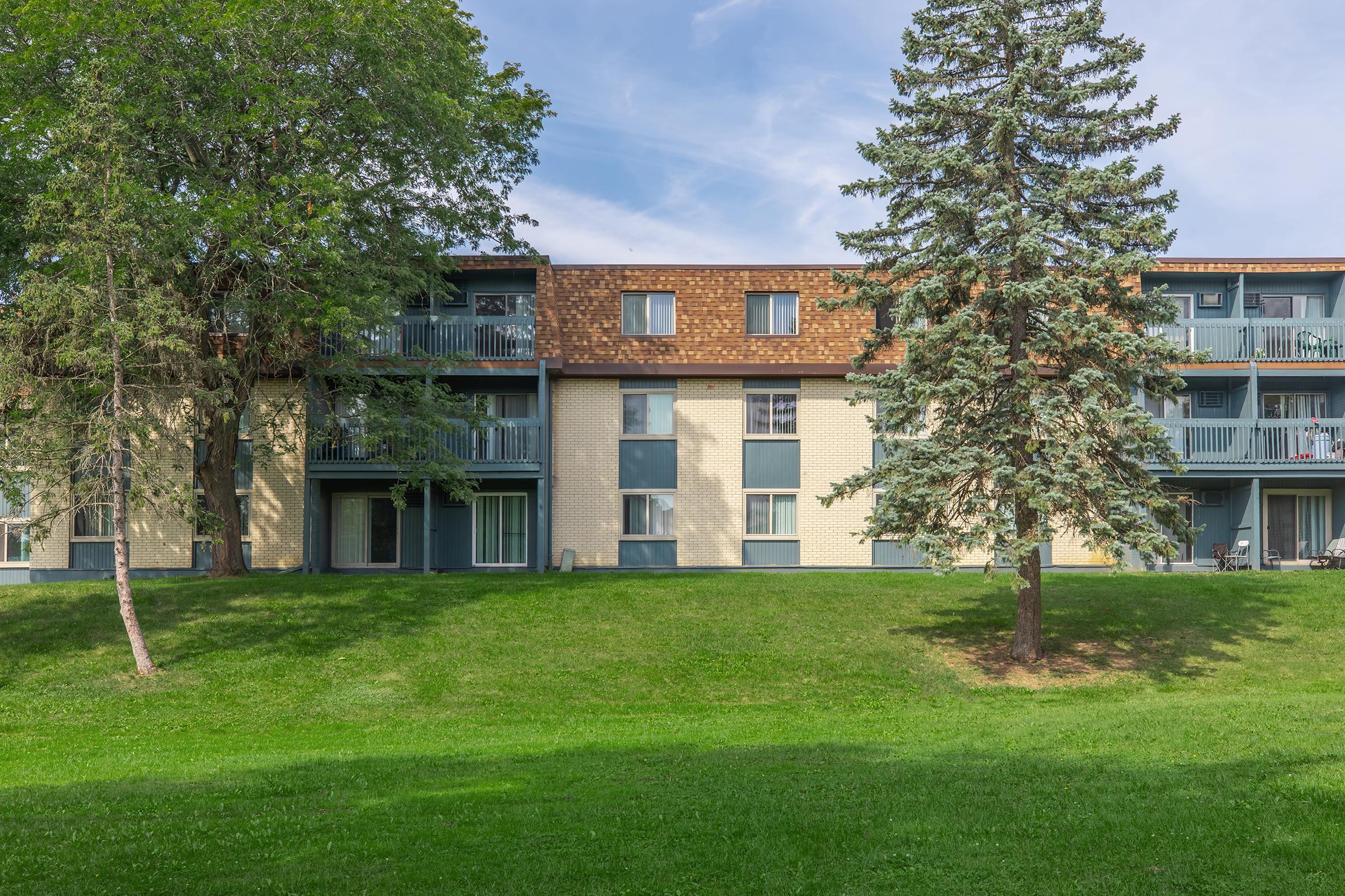 A multi-story residential building with a grassy area in front. The structure has a light-colored brick façade and wooden accents on the upper floor. Large windows and balconies are visible, surrounded by trees and greenery, creating a peaceful, suburban atmosphere.