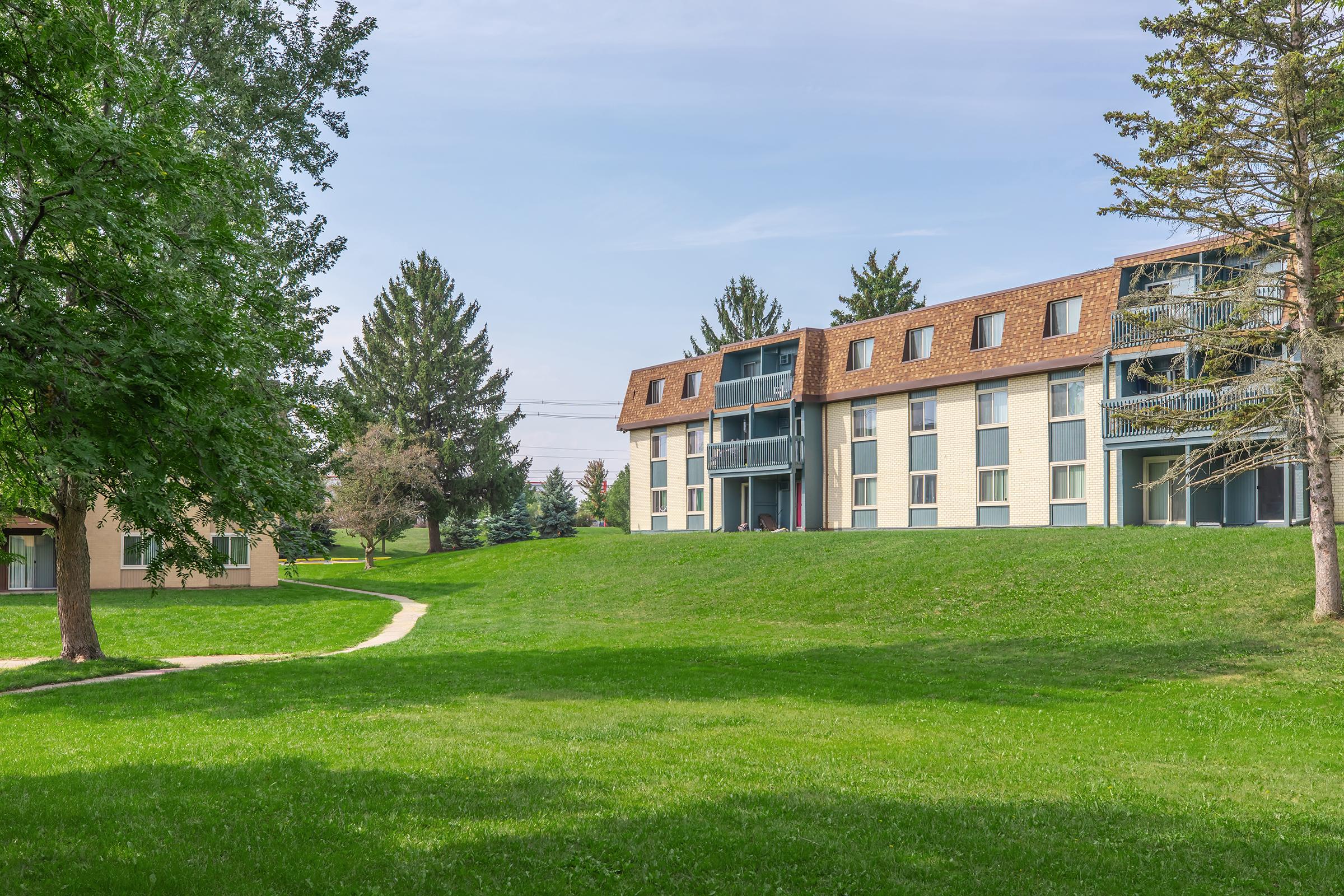 A grassy area featuring two buildings. The main building is three stories tall with a brown roof and multiple balconies. Trees and shrubs surround the buildings, and a walking path leads through the green lawn. The sky is clear with some clouds.