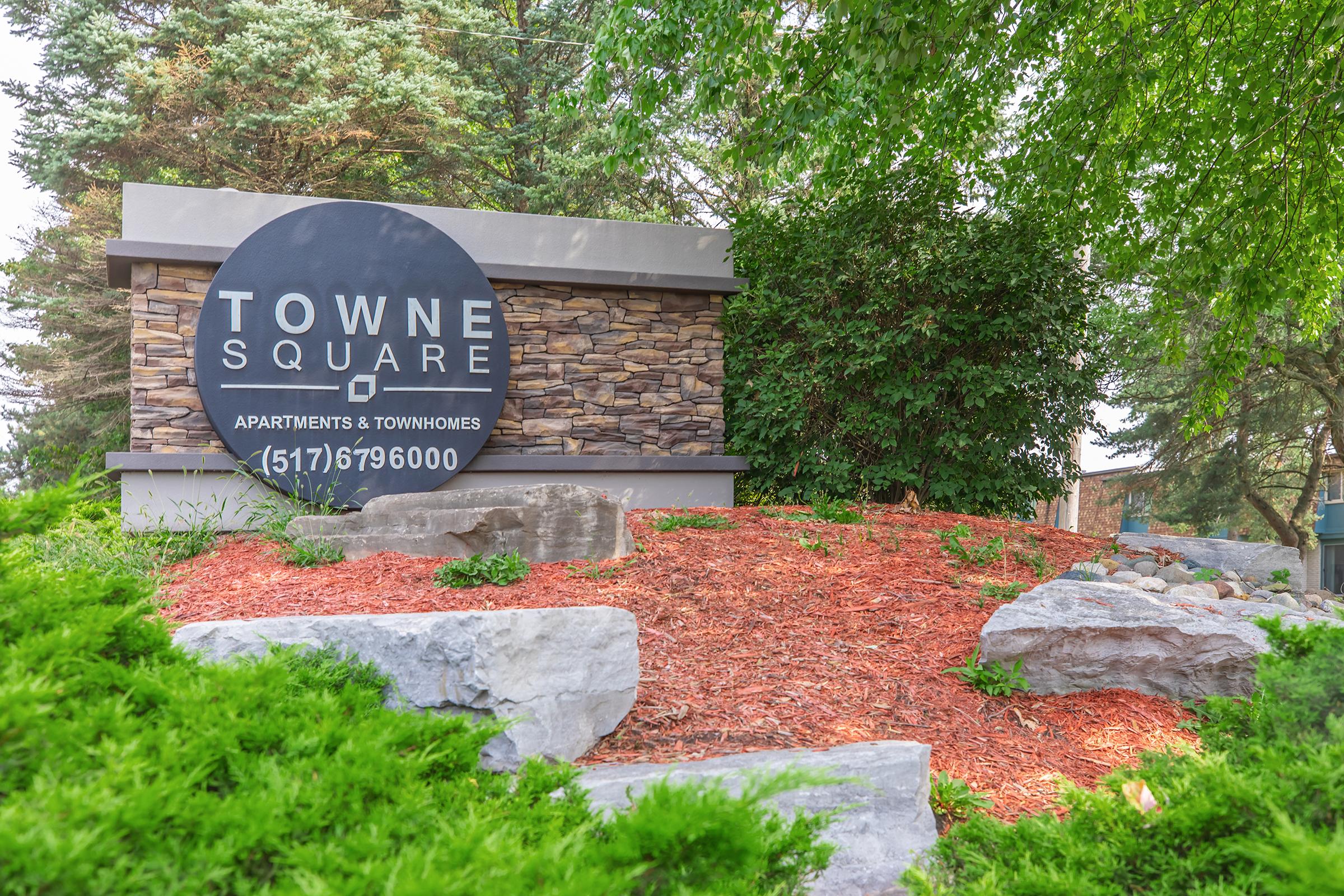 Sign for Towne Square Apartments & Townhomes, featuring a black circular logo with white text. The sign is surrounded by landscaped greenery, rocks, and mulch, indicating an inviting residential area. The contact number is displayed as (517) 679-6000.