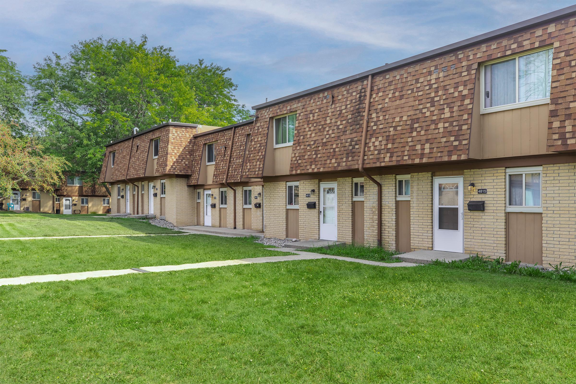 A row of residential townhouses with brown shingle roofs and beige brick walls, set in a green lawn area. The buildings feature white doors and small pathways leading to each unit, with a backdrop of trees in the distance. The scene is bright and well-maintained, suggesting a quiet residential neighborhood.