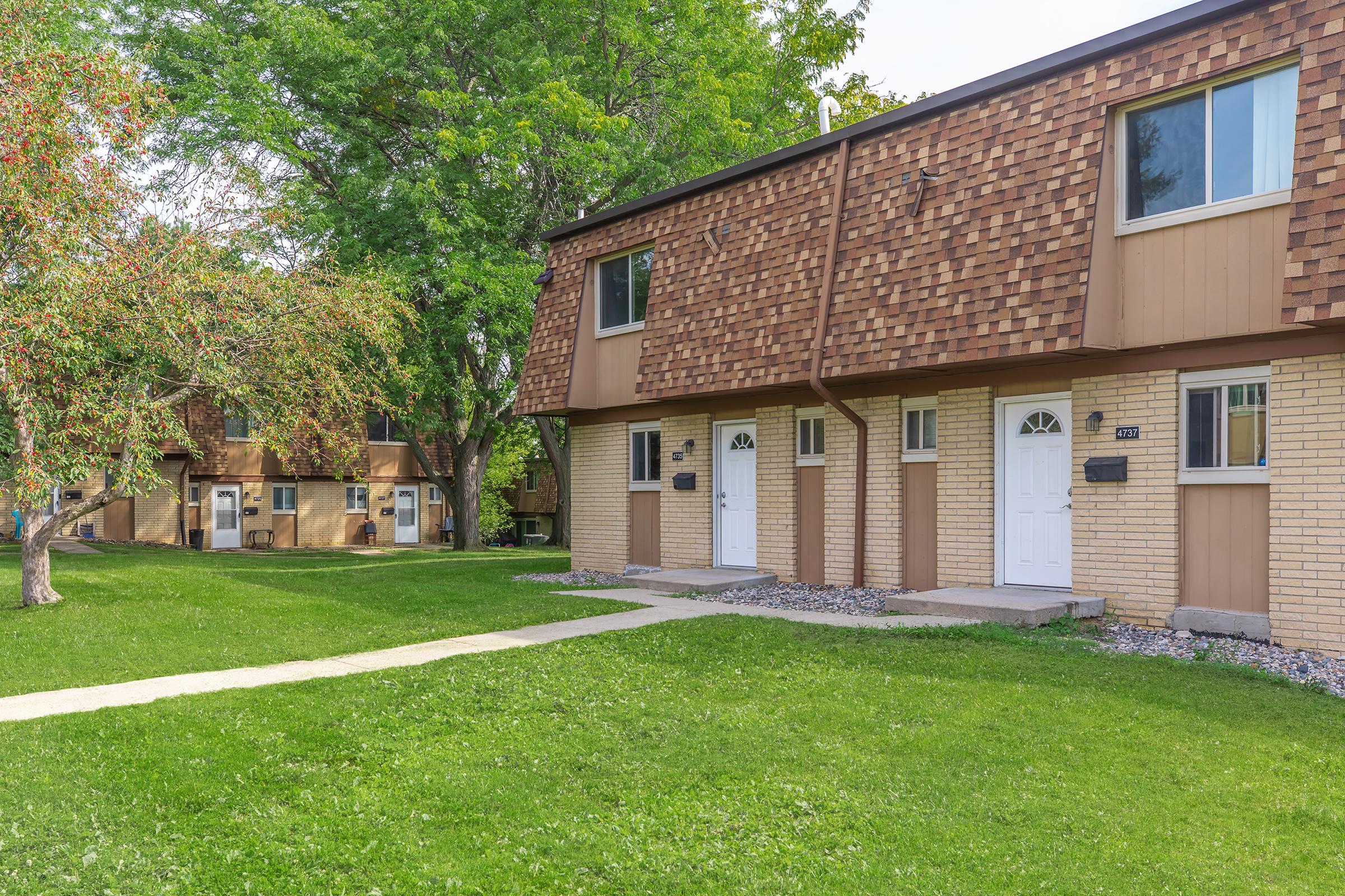 A row of townhouses with two-story structures featuring a mix of brick and wood siding. Each townhouse has a distinct front door and a small pathway leading to it, surrounded by well-kept grass. In the background, additional similar townhouses and trees can be seen, providing a tranquil residential setting.