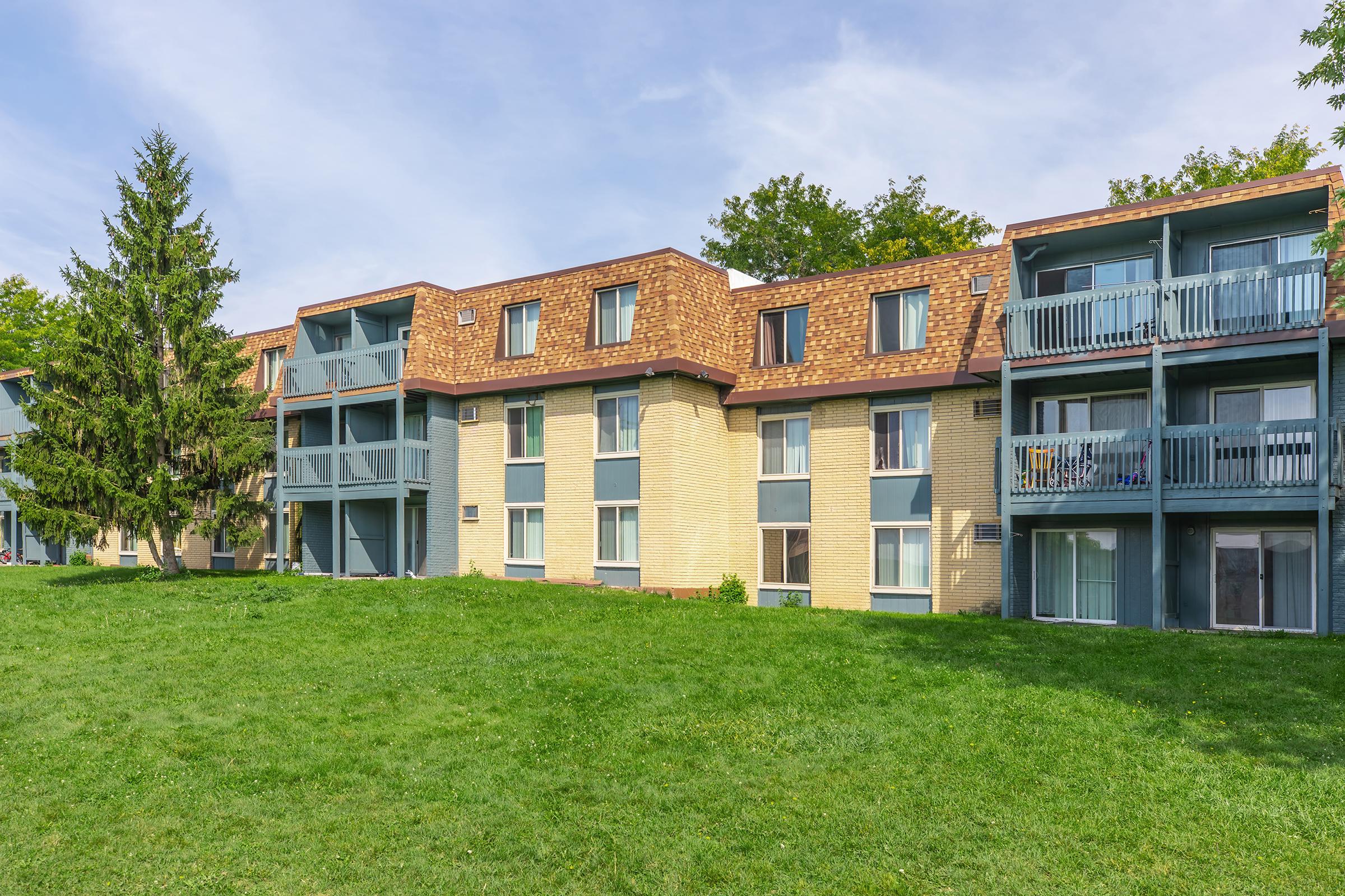 A multi-story residential building with a mix of brick and siding. The structure features several balconies and large windows, surrounded by a green grassy area and trees in the background. The sky is clear with a few clouds, indicating a bright day.
