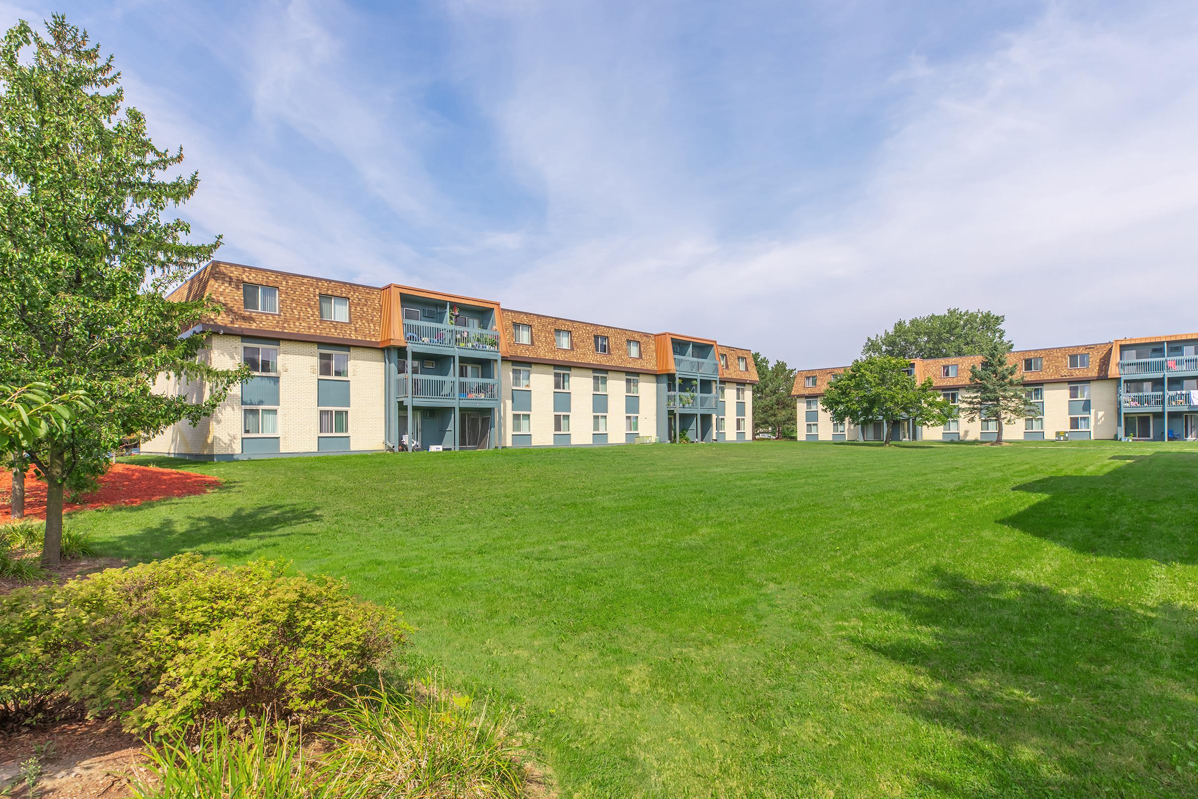 A view of a multi-story apartment complex surrounded by lush green grass and trees. The building features a light exterior with brown roof accents, and balconies on the upper floors. Clear blue skies and a well-maintained landscape enhance the tranquil atmosphere.