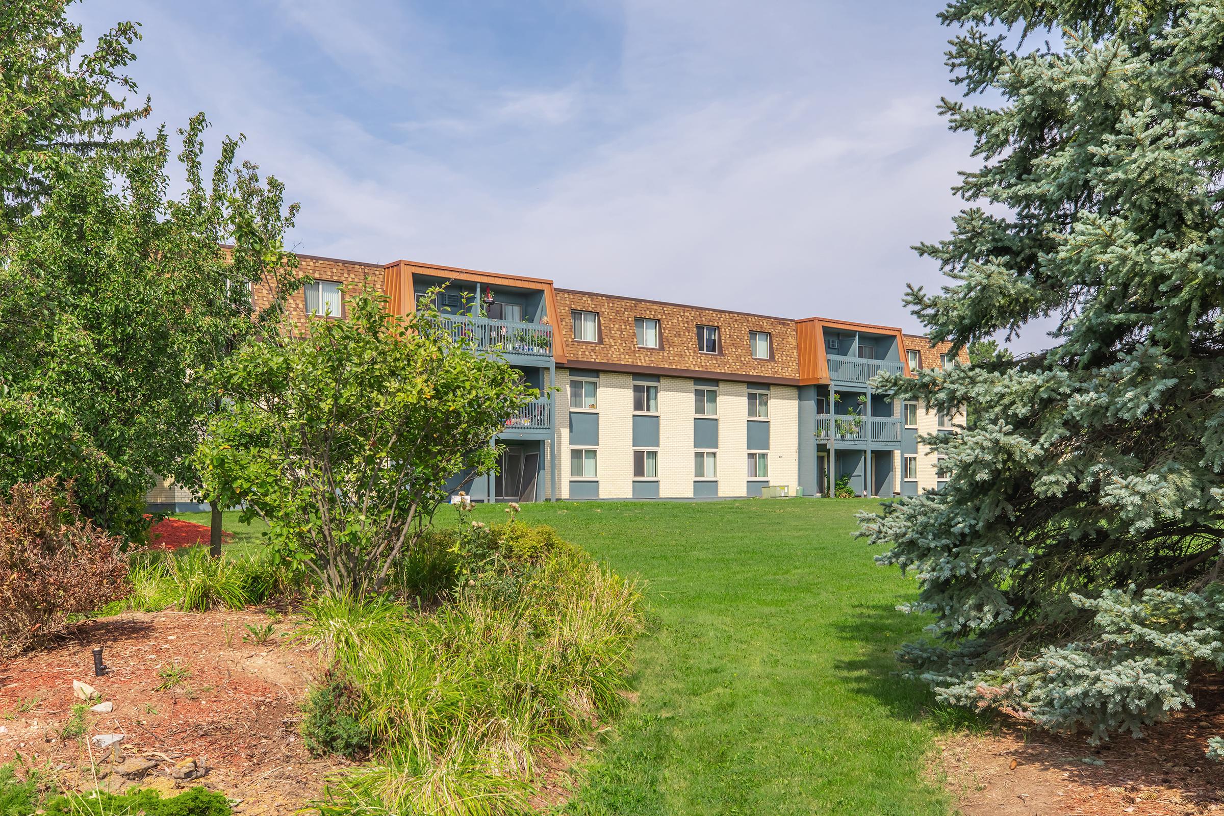 A low-rise apartment building with a mix of colors, surrounded by greenery. The structure features balconies on the upper floors, with a well-maintained lawn and various shrubs in the foreground. The sky is partly cloudy, contributing to a pleasant outdoor setting.