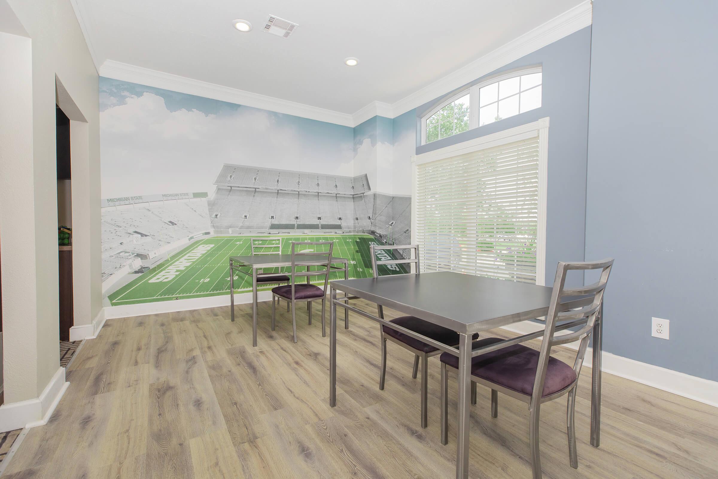 A modern dining area featuring a dark wood table surrounded by four silver chairs. The wall displays a large mural of a football stadium, creating a sporty ambiance. Natural light streams through the window, and the flooring is a light wood finish, adding warmth to the space.