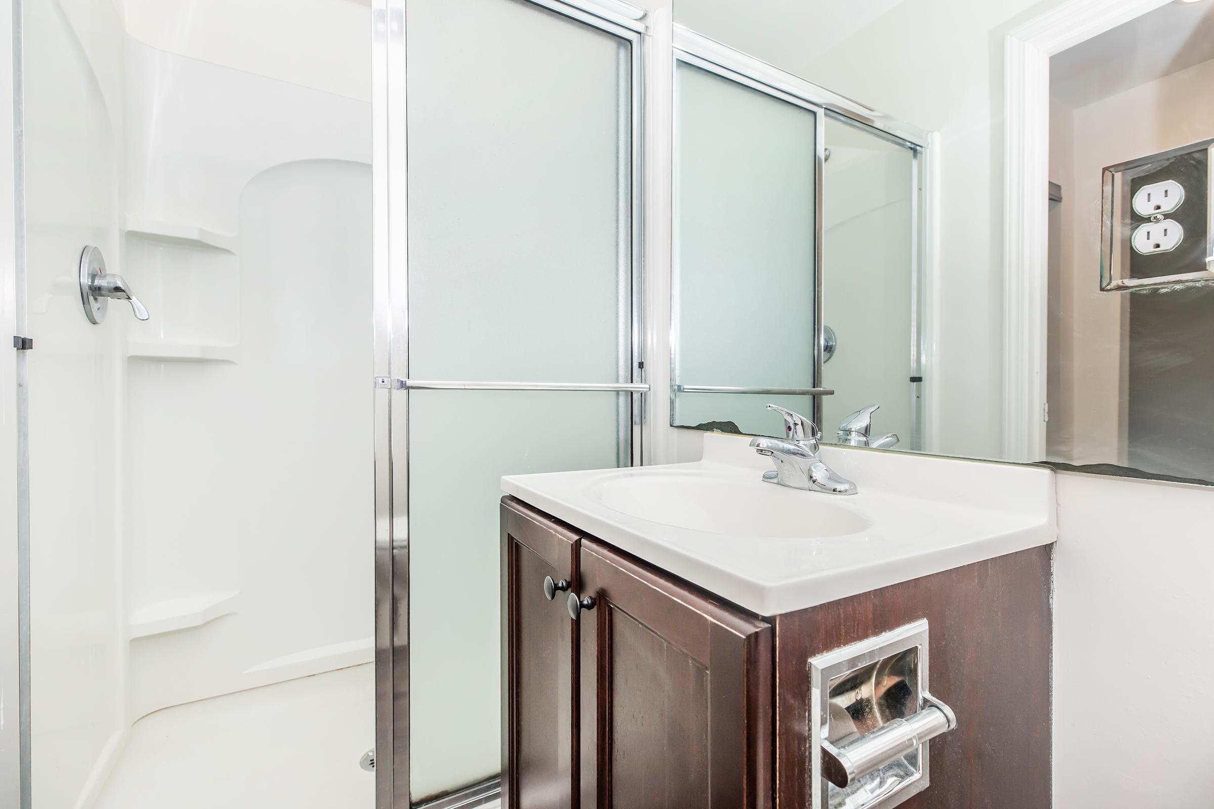 A modern bathroom featuring a shower stall with glass doors, a white sink with a chrome faucet, and dark wood cabinetry. The walls are painted in light colors, and there is a mirror along one side. Electric outlets are visible near the sink area.