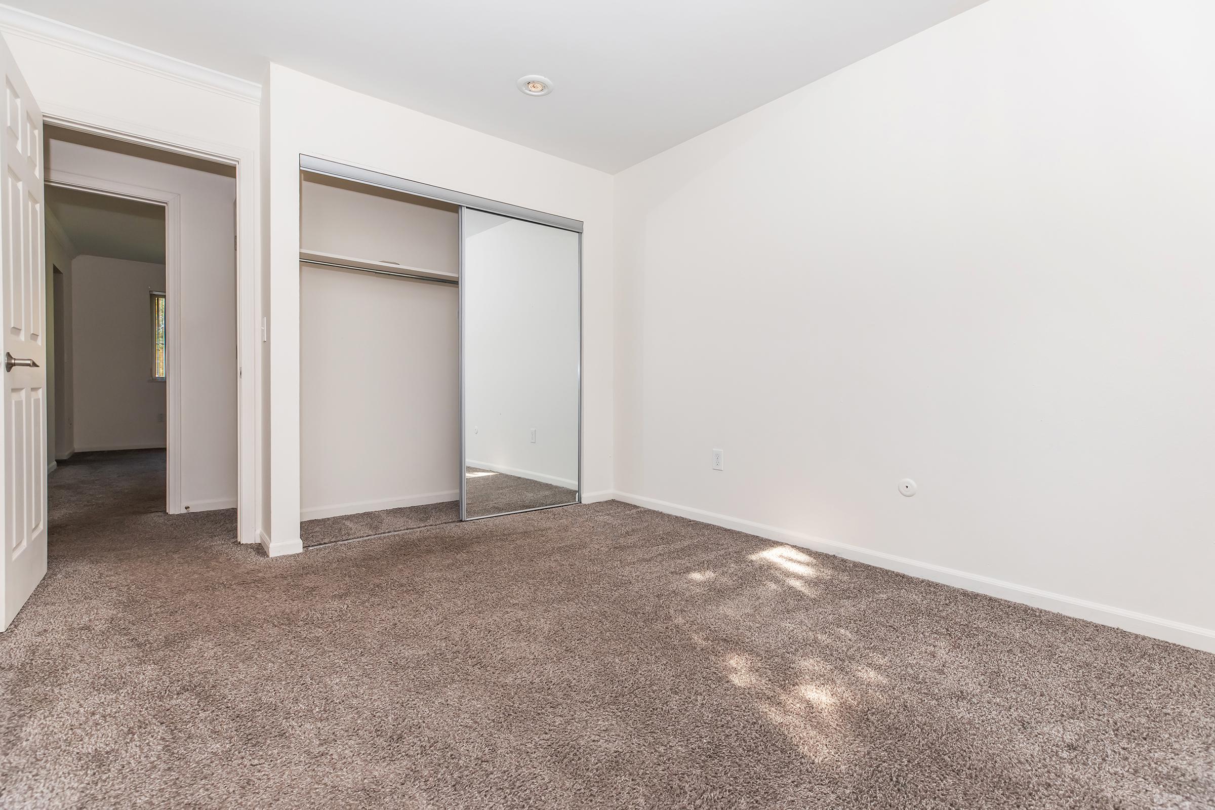 A spacious, empty room with light beige walls and a large mirror sliding closet. The floor is covered in plush carpet. A doorway leads to another room, and a window allows natural light to enter, creating a bright and airy atmosphere.