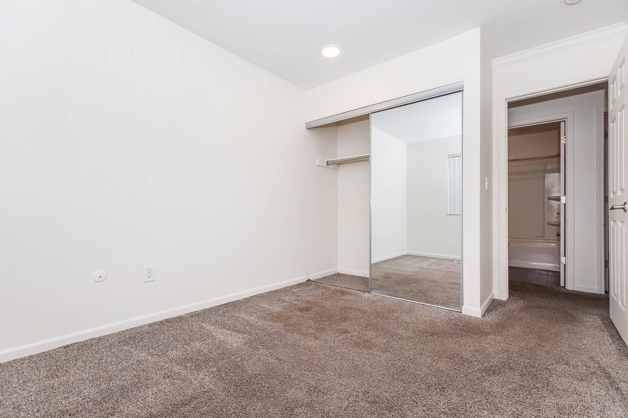 A vacant bedroom featuring light-colored walls and brown carpet. There is a closet with mirrored doors on one side and a bathroom entrance visible in the background, suggesting a tidy and well-maintained space ready for new occupants. Natural light enters through a window, creating a welcoming atmosphere.