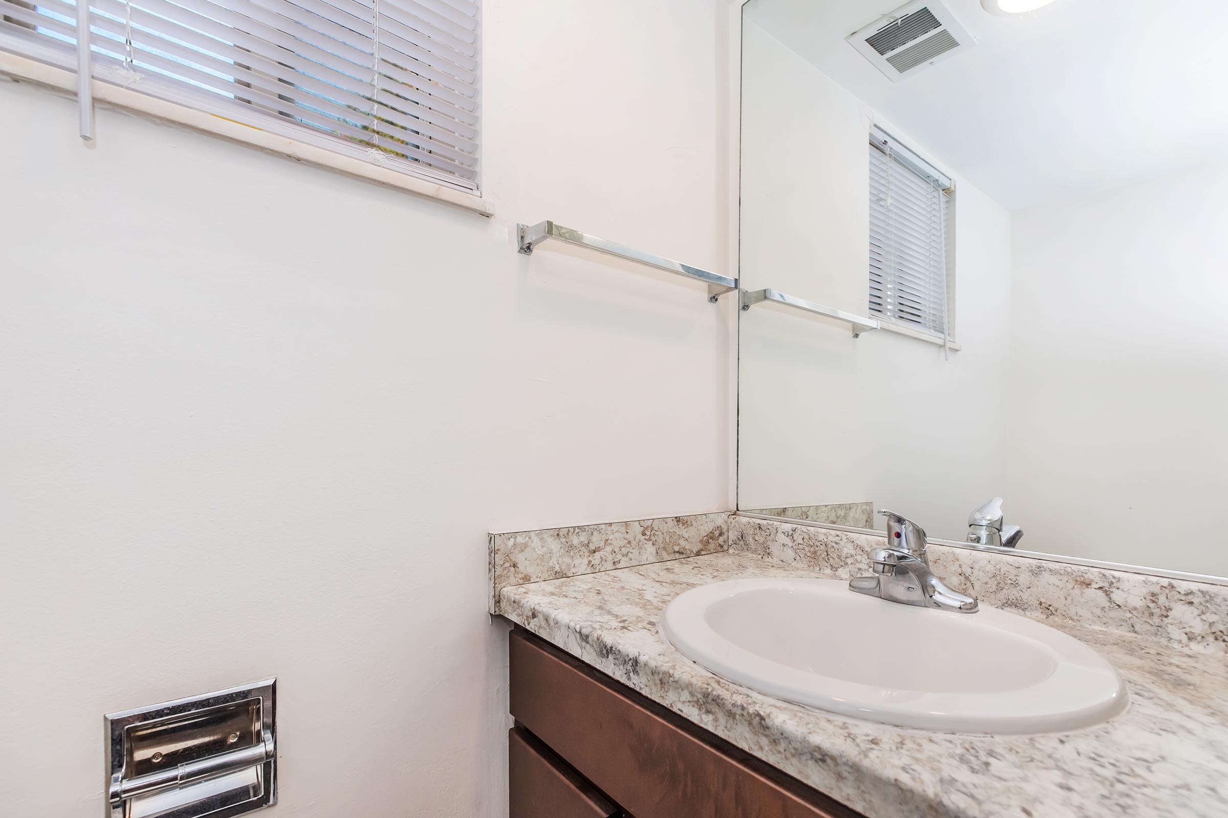A clean bathroom countertop featuring a round sink with a chrome faucet. The counter is made of light-colored granite. Above it, there is a mirror reflecting the room. A towel bar and a window with blinds are also visible, contributing to a bright and tidy atmosphere.