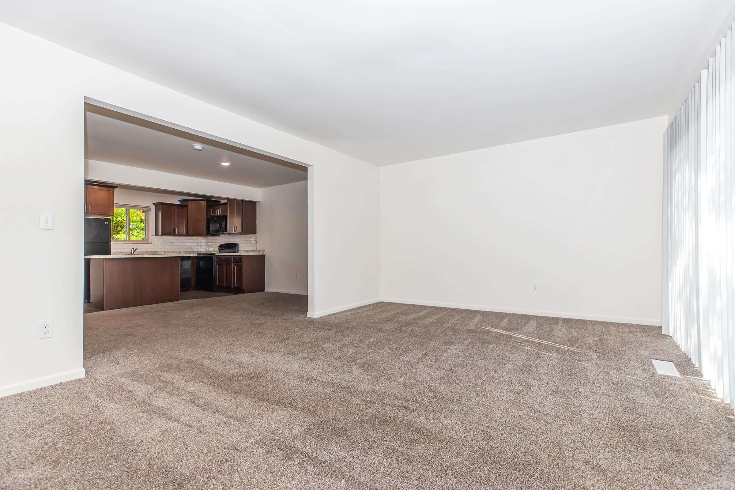 A spacious, empty living room with light beige carpet and white walls. An open doorway leads to a modern kitchen featuring dark wood cabinetry and black appliances. Natural light fills the space, highlighting the airy atmosphere.
