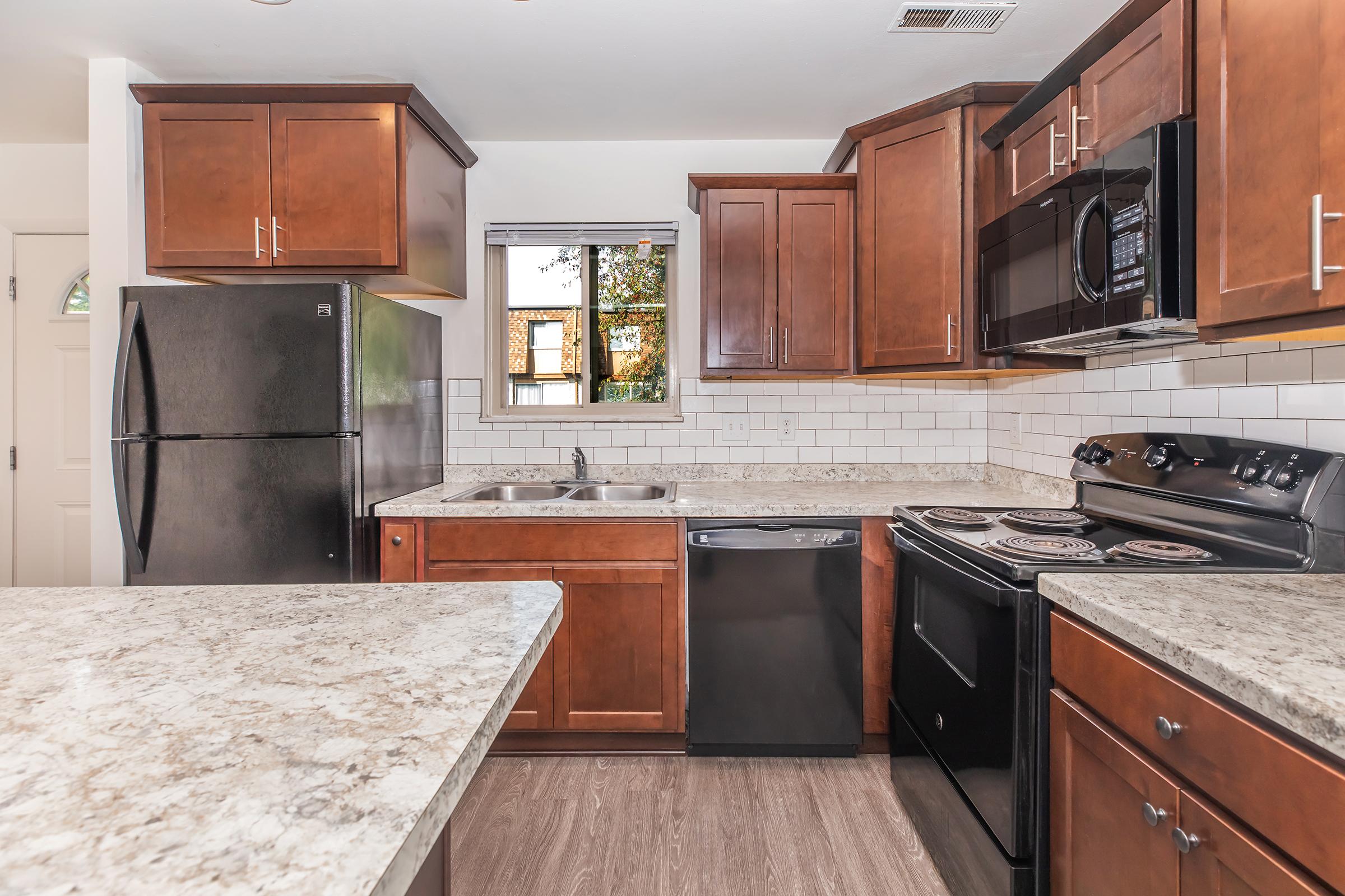 A modern kitchen featuring wooden cabinets, a stainless steel sink, and granite countertops. Appliances include a black refrigerator, microwave, and dishwasher, along with a black stove and oven. The kitchen has a window that lets in natural light, enhancing the warm and inviting atmosphere.