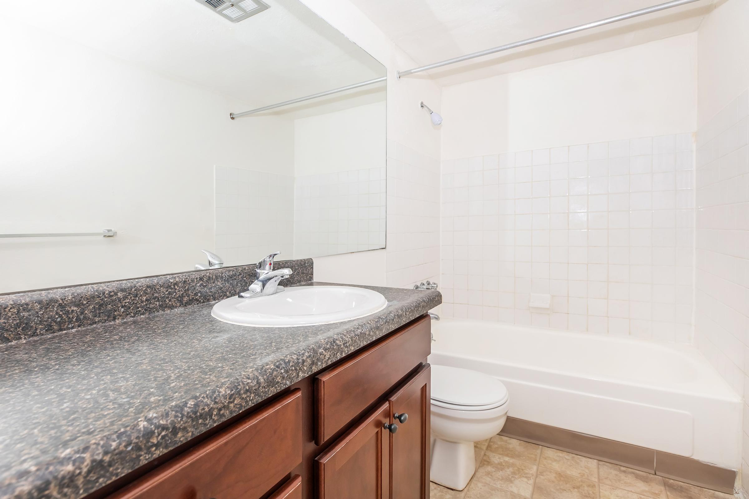 A clean bathroom featuring a granite countertop with a sink, a mirrored wall, a bathtub with white tiled walls, and a toilet. The floor is tiled, and the overall design has a modern, simple aesthetic.