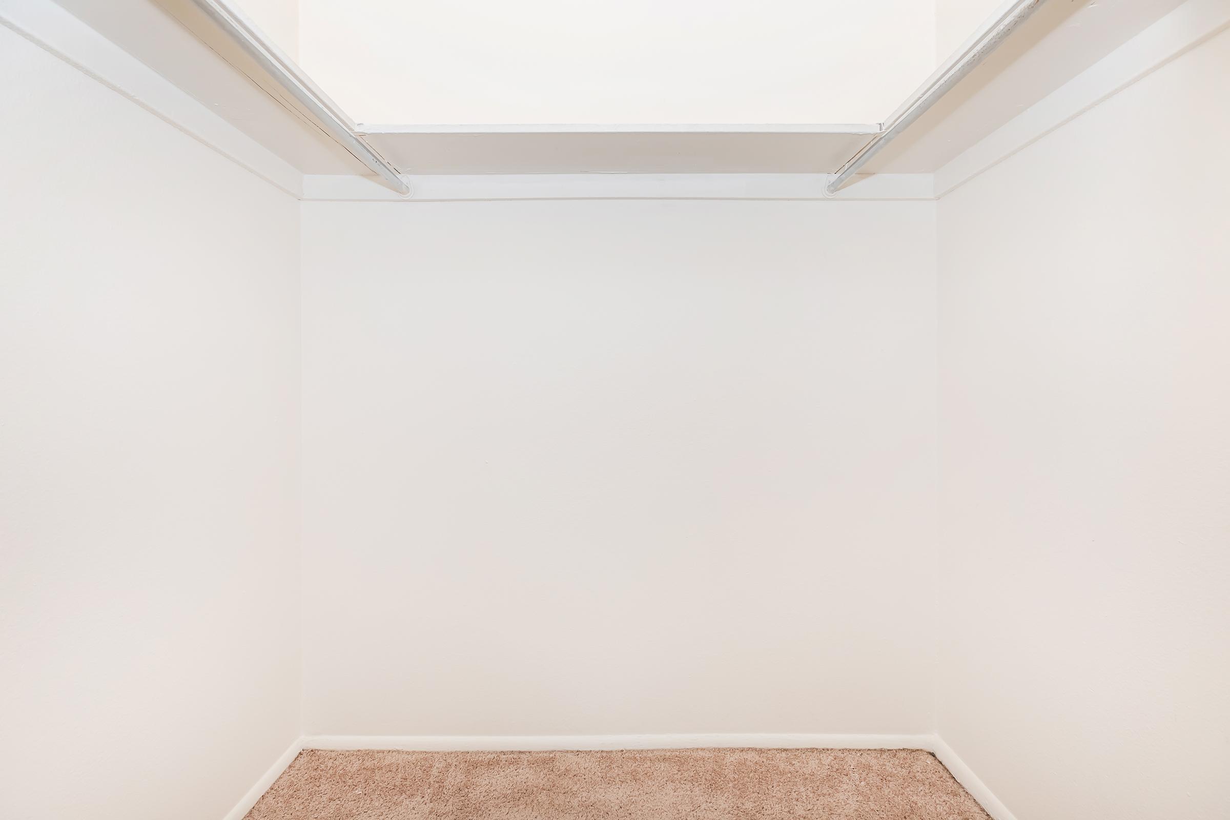 An empty closet with white walls and a carpeted floor. The ceiling is visible through an opening above, and there are no items or shelves inside the closet, creating a spacious but minimalist appearance.