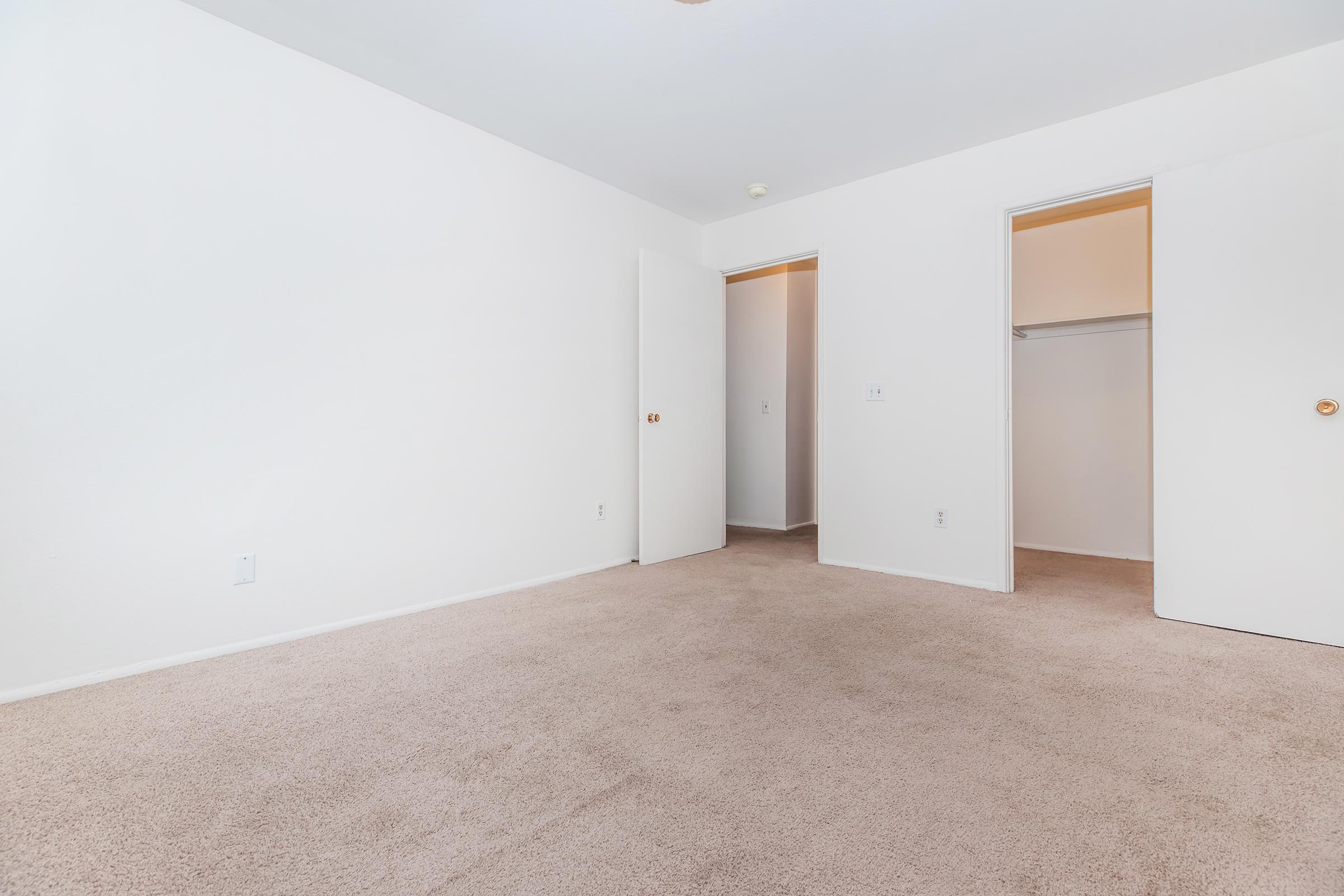 A vacant room with beige carpet and plain white walls. The room features two open doorways leading to closets and additional spaces, with no furniture or decorations present. Natural light illuminates the area, creating a bright and airy atmosphere.