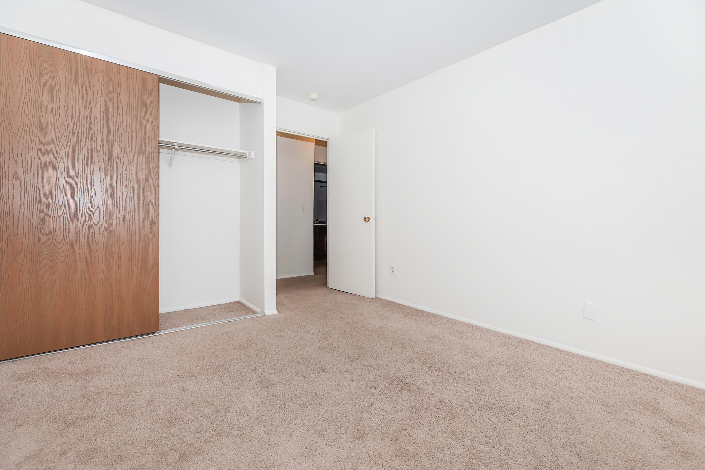 A vacant room with beige carpet, white walls, and a wooden closet door. The space features a hanging rack inside the closet and a partially open door leading to another room. The overall atmosphere is bright and neutral, creating a simple and uncluttered environment.