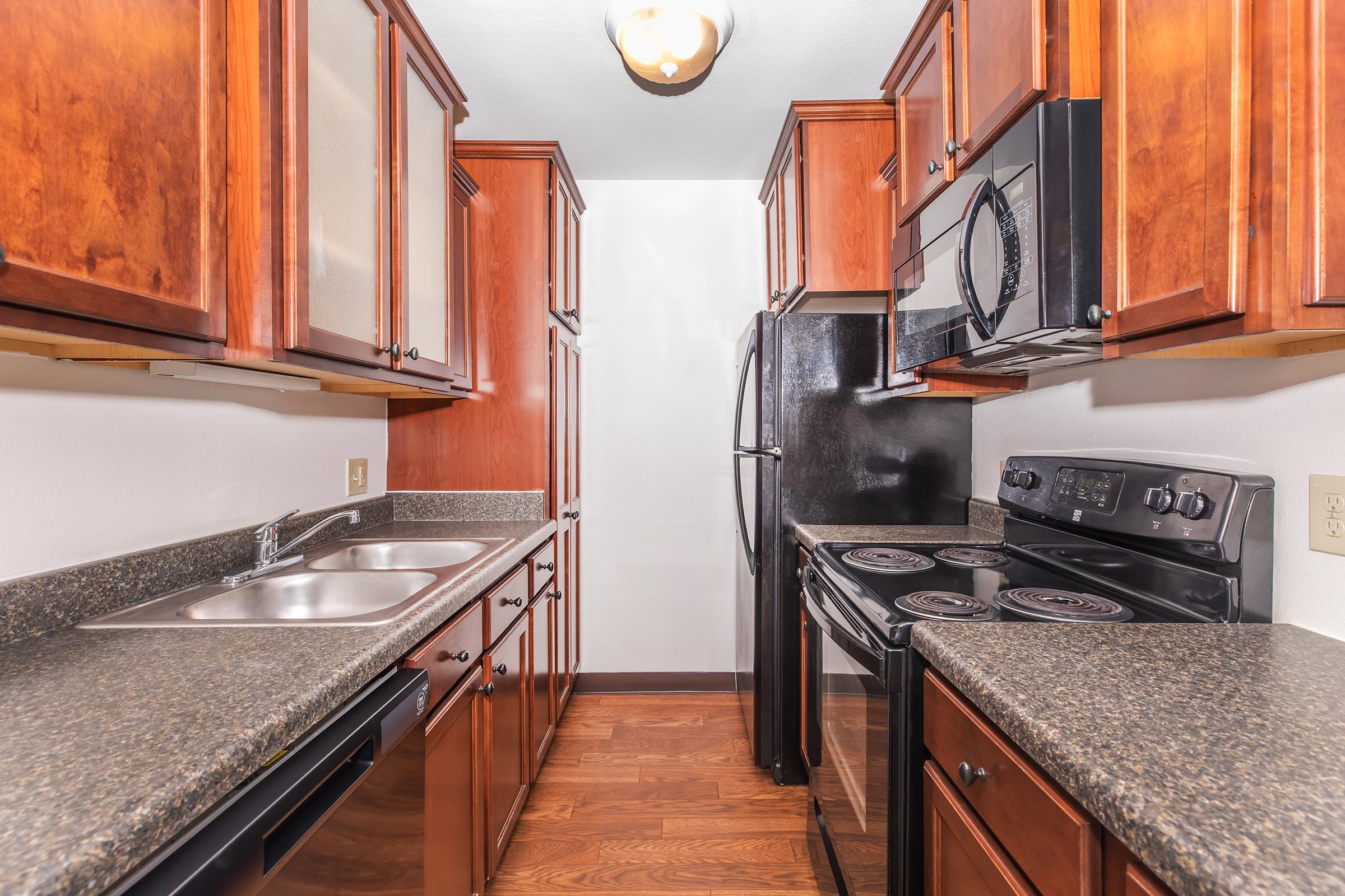 A modern kitchen featuring dark wood cabinets, stainless steel appliances, and black countertops. The space includes a double sink, electric stove, and an over-the-range microwave, with ample cabinet storage. The flooring is a warm brown, complementing the cabinetry. Natural light from a ceiling fixture illuminates the area.
