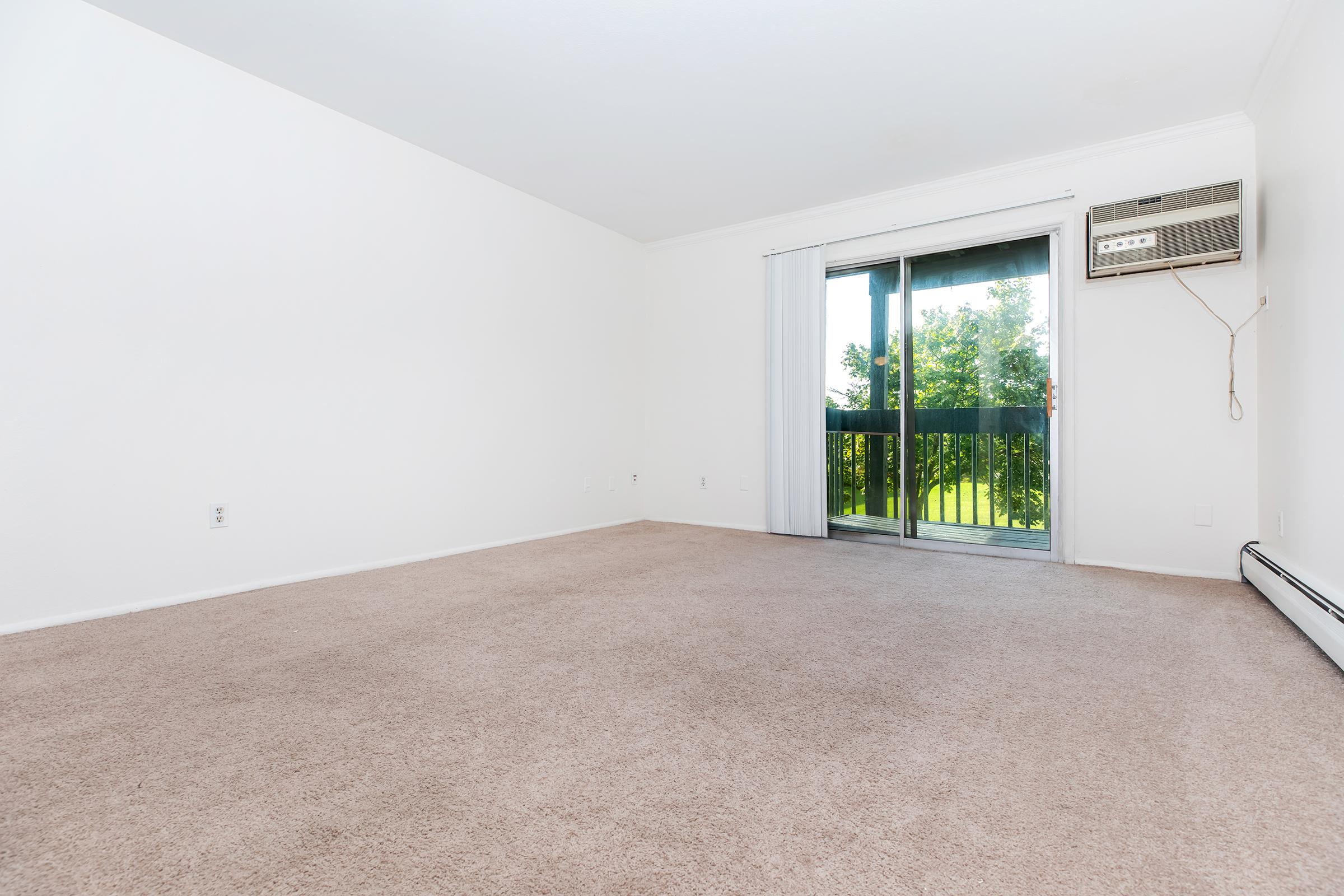 A spacious, minimally furnished room with light beige carpet and white walls. A large sliding glass door leads to a balcony with green foliage visible outside. An air conditioning unit is mounted on the wall near the door, and the overall ambiance is bright and airy, suggesting potential for personalization.