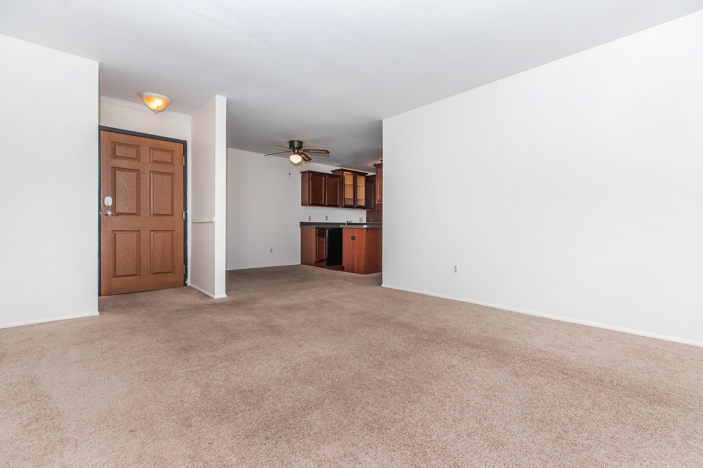 Spacious, empty living room with beige carpet, a single ceiling fan, and light-colored walls. A wooden front door with a peephole is visible on the left side. In the background, a kitchen area features dark wooden cabinetry, but there are no visible appliances or furnishings.