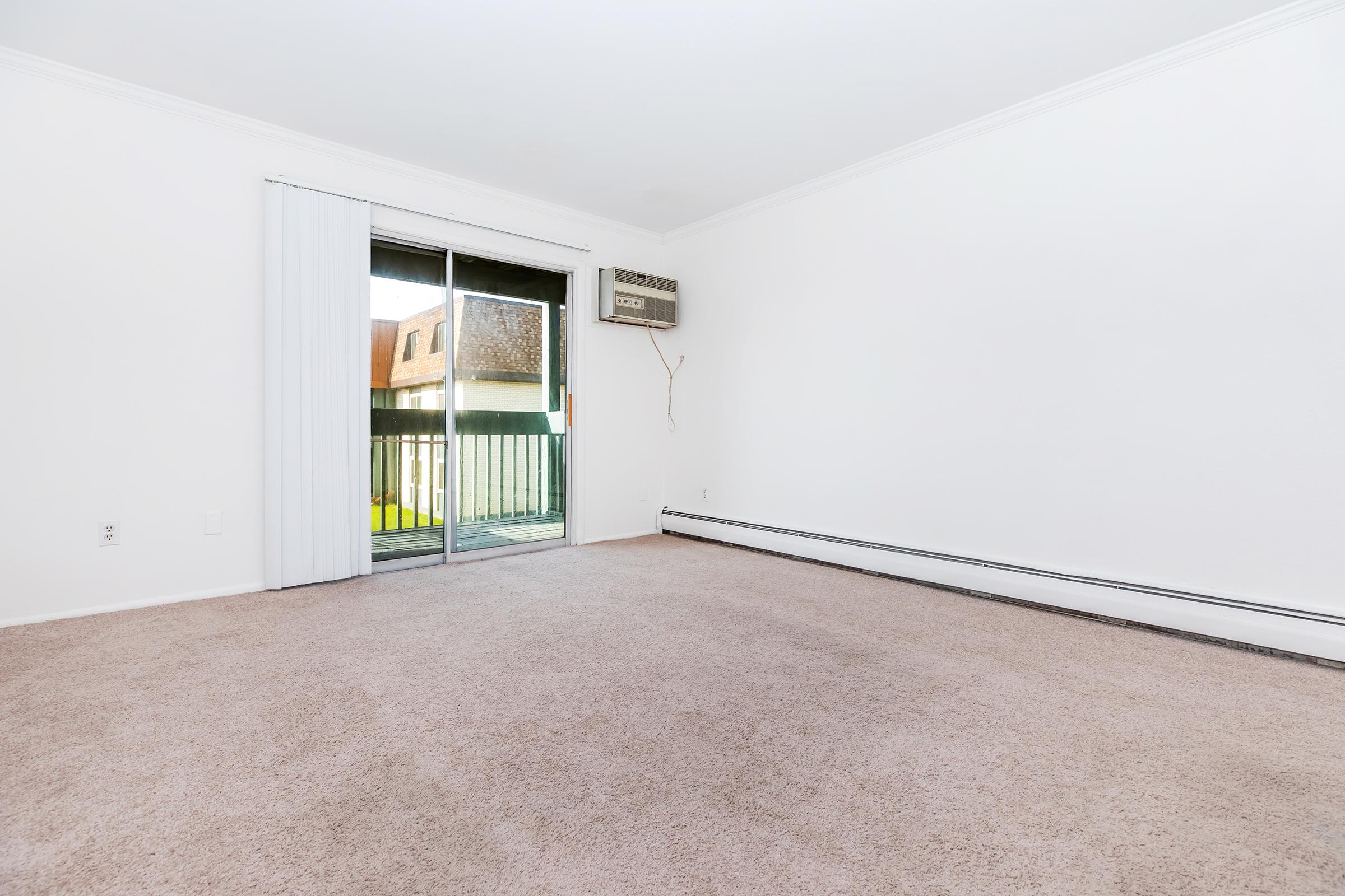 Empty room with light beige carpet and white walls. A sliding glass door leads to a small balcony, and an air conditioning unit is mounted on the wall. The space is bright and uncluttered, suggesting potential for personalization.
