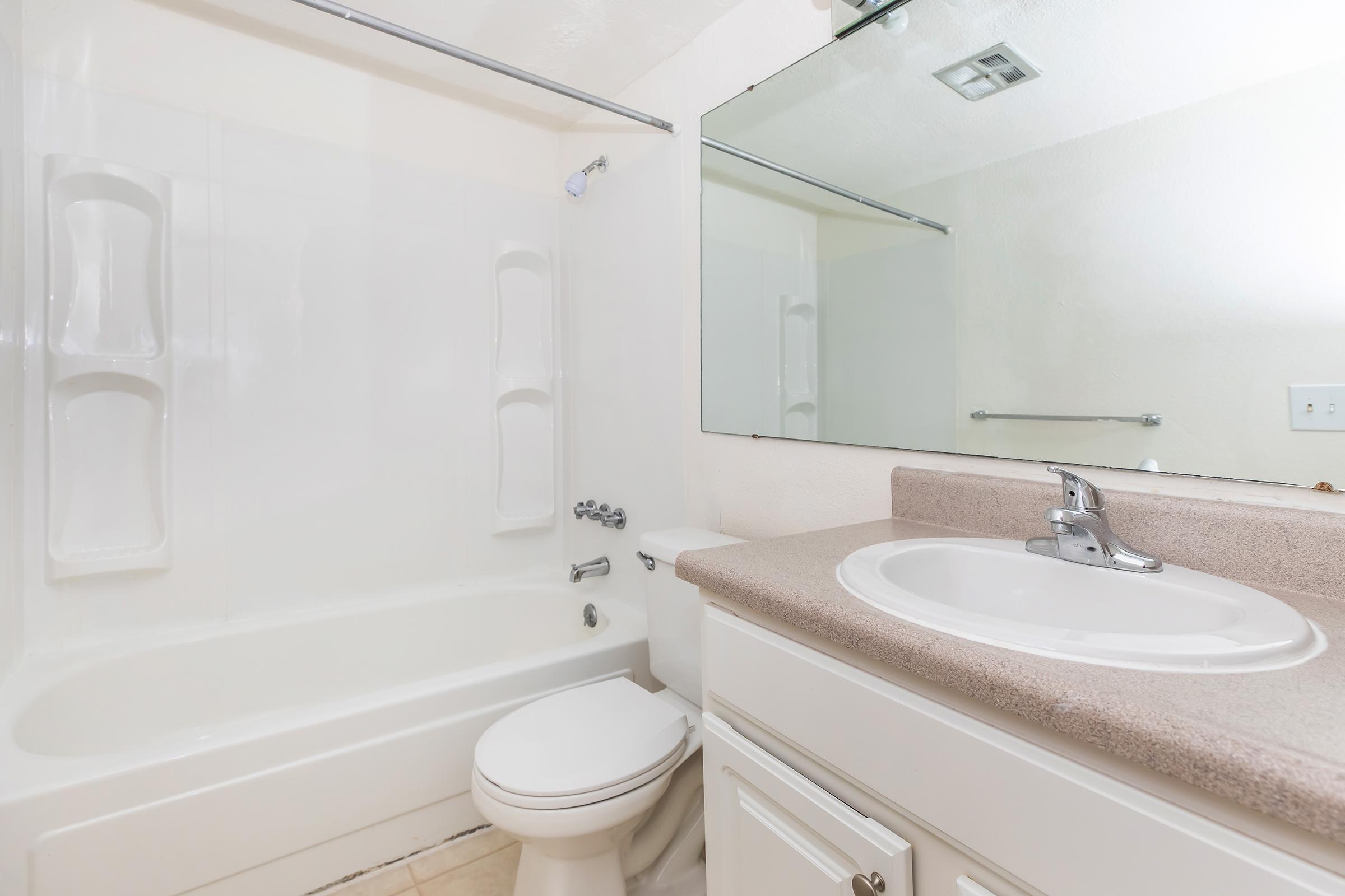 A clean and modern bathroom featuring a white bathtub with an integrated shower, a single sink vanity with a light-colored countertop, a large mirror above the sink, and neutral-colored walls. The space is well-lit and neatly organized, providing a contemporary feel.