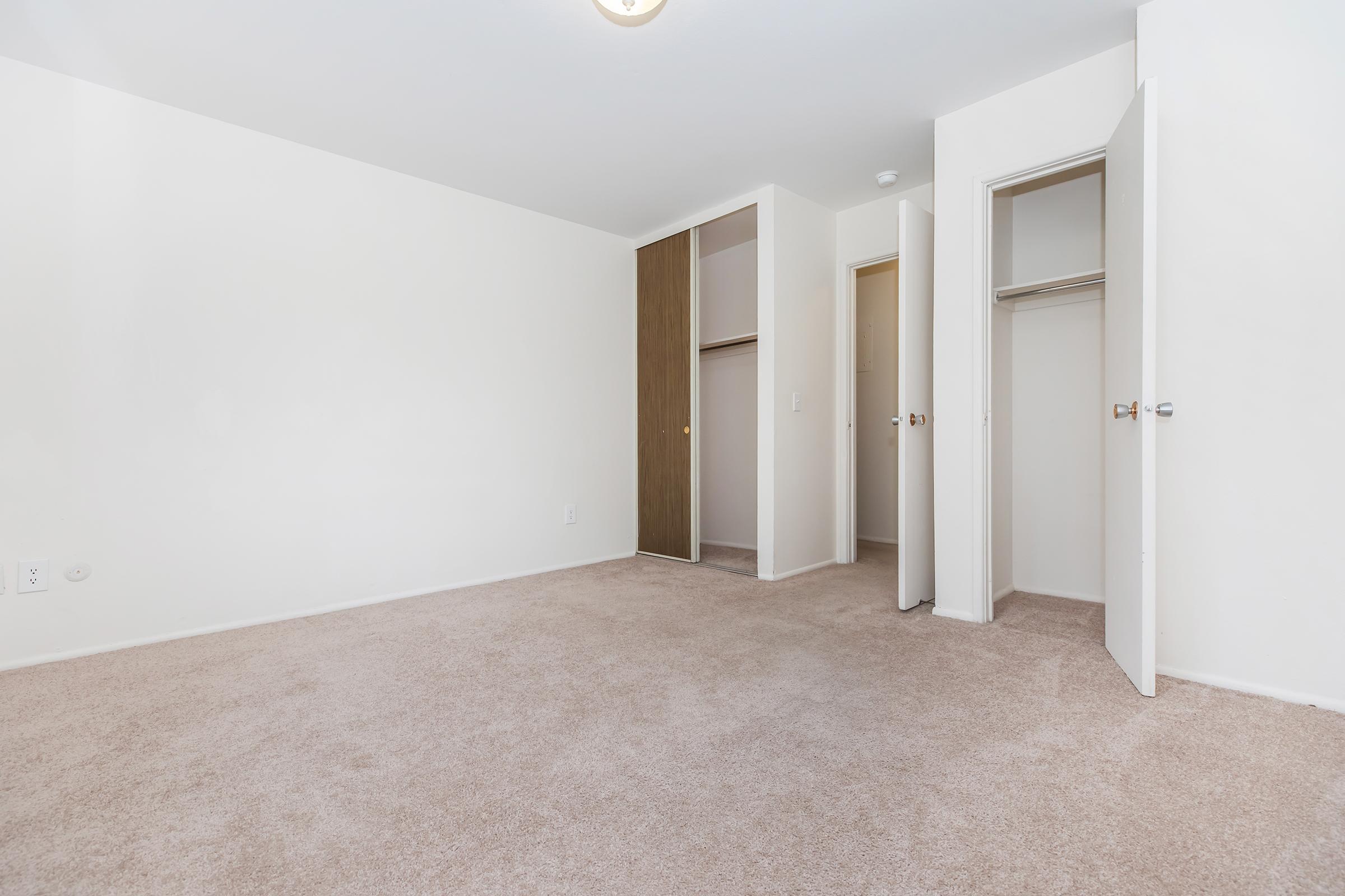 A spacious, empty room with light-colored walls and beige carpet. Two open closets with wooden doors on the right side. A ceiling light fixture illuminates the space. The room appears bright and uncluttered, suitable for various uses such as a bedroom or office.