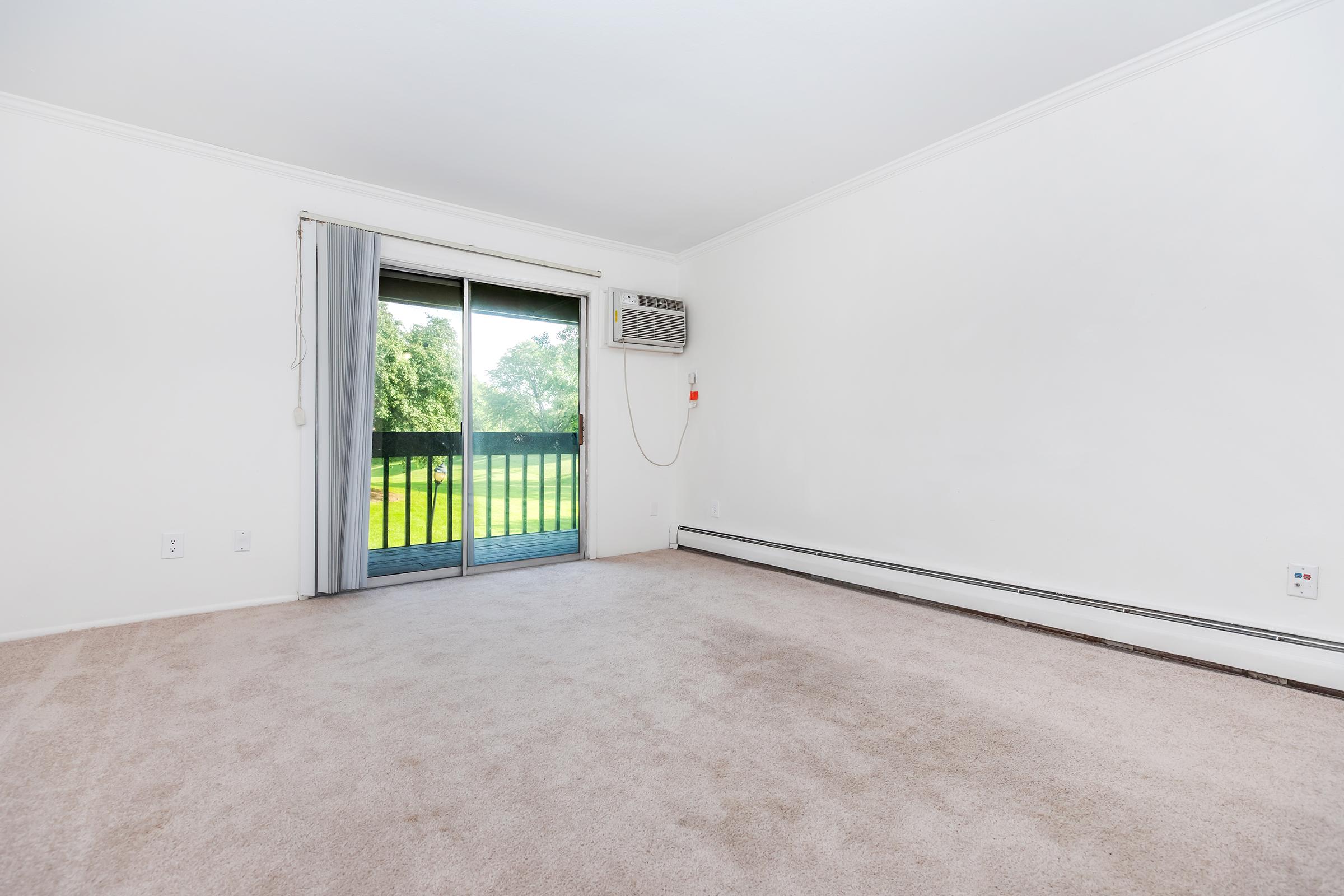 A spacious, empty living room with light-colored carpet, a sliding glass door leading to a balcony, and an air conditioning unit mounted on the wall. The walls are painted white, and the space is filled with natural light, creating a bright and airy atmosphere.