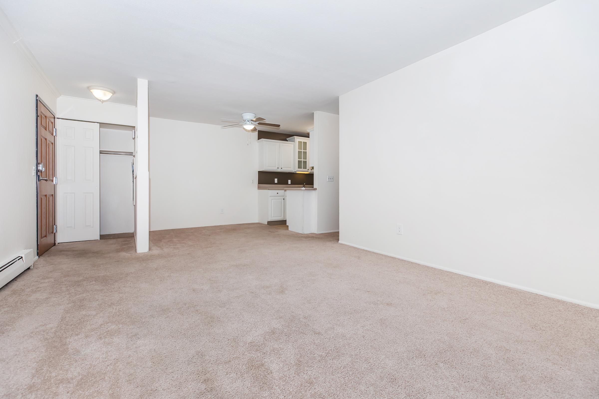 A spacious, empty living area with beige carpet and white walls. To the left, there is a closet with wooden doors. In the background, a small kitchen area with cabinetry is visible. A ceiling fan hangs overhead, and there is a light fixture in the ceiling. The overall ambiance is bright and inviting.