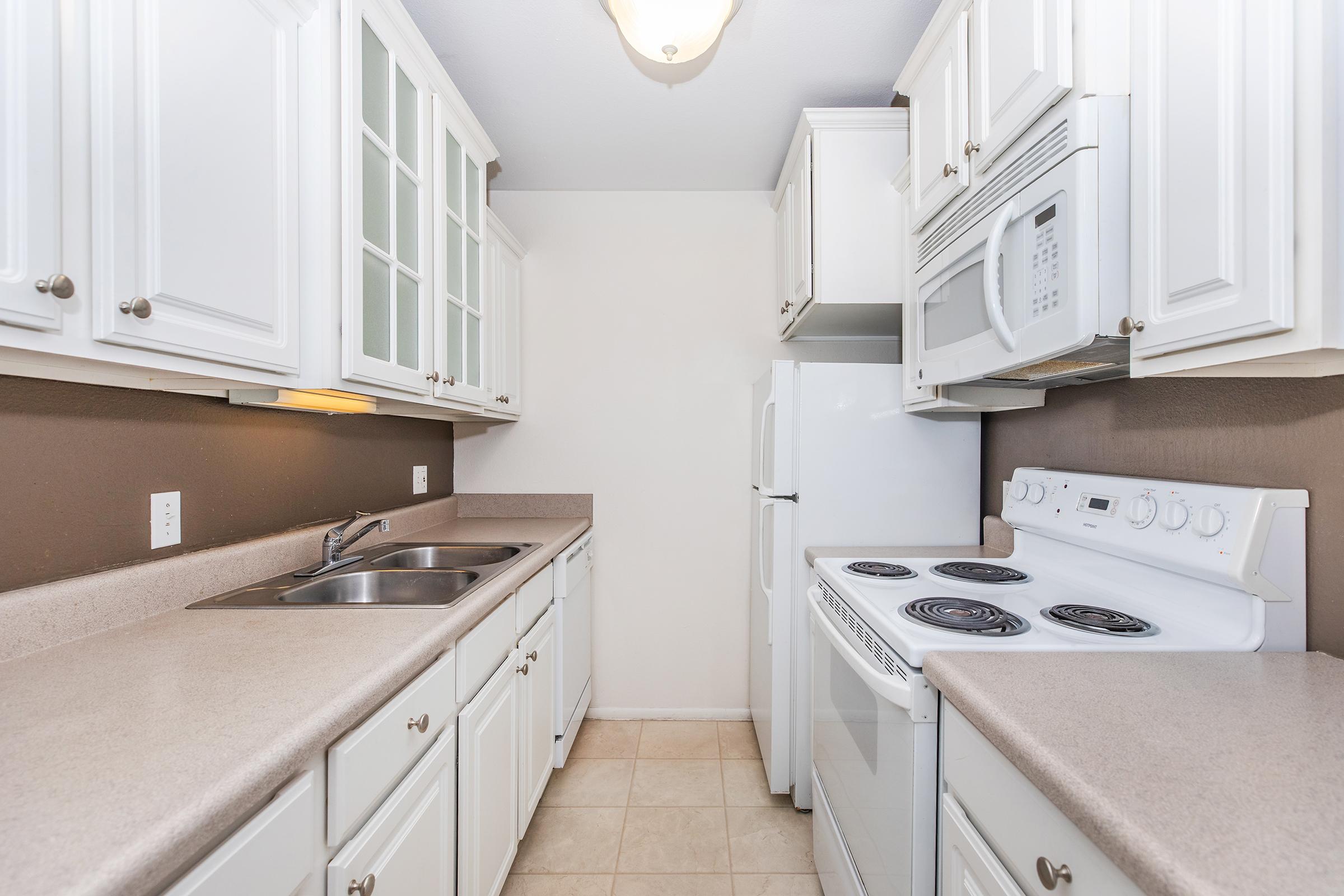 A modern kitchen featuring white cabinetry, a double sink, stainless steel appliances, and a light-colored countertop. The layout includes an oven, stove, microwave, and refrigerator, with ample storage and a clean, simple design. The walls are painted in a neutral tone, creating a bright and inviting atmosphere.