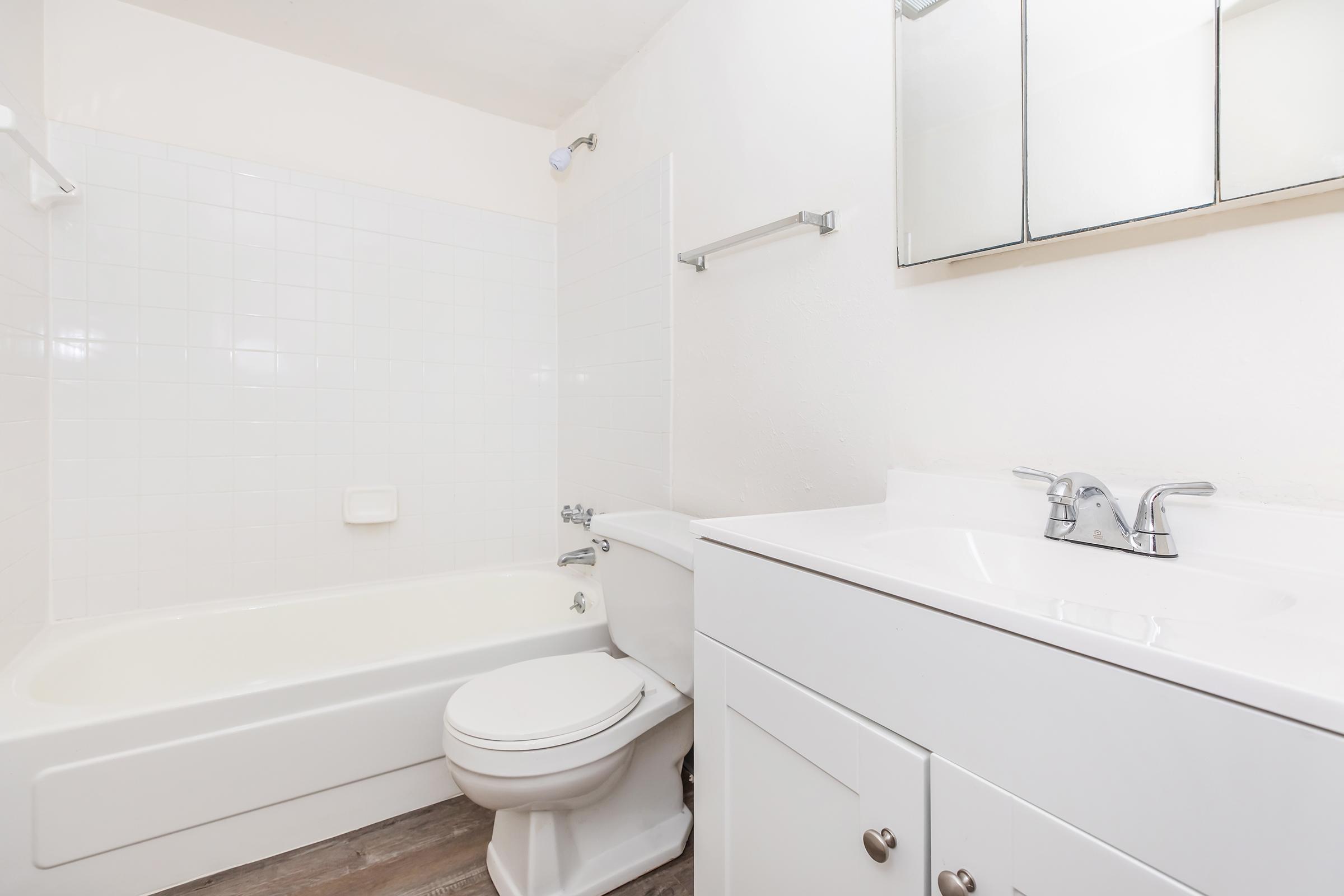 A clean and modern bathroom featuring a white bathtub with a shower, a white toilet, and a white sink with a mirror above it. The walls are tiled in light colors, and the floor is a neutral tone, creating a bright and inviting space.