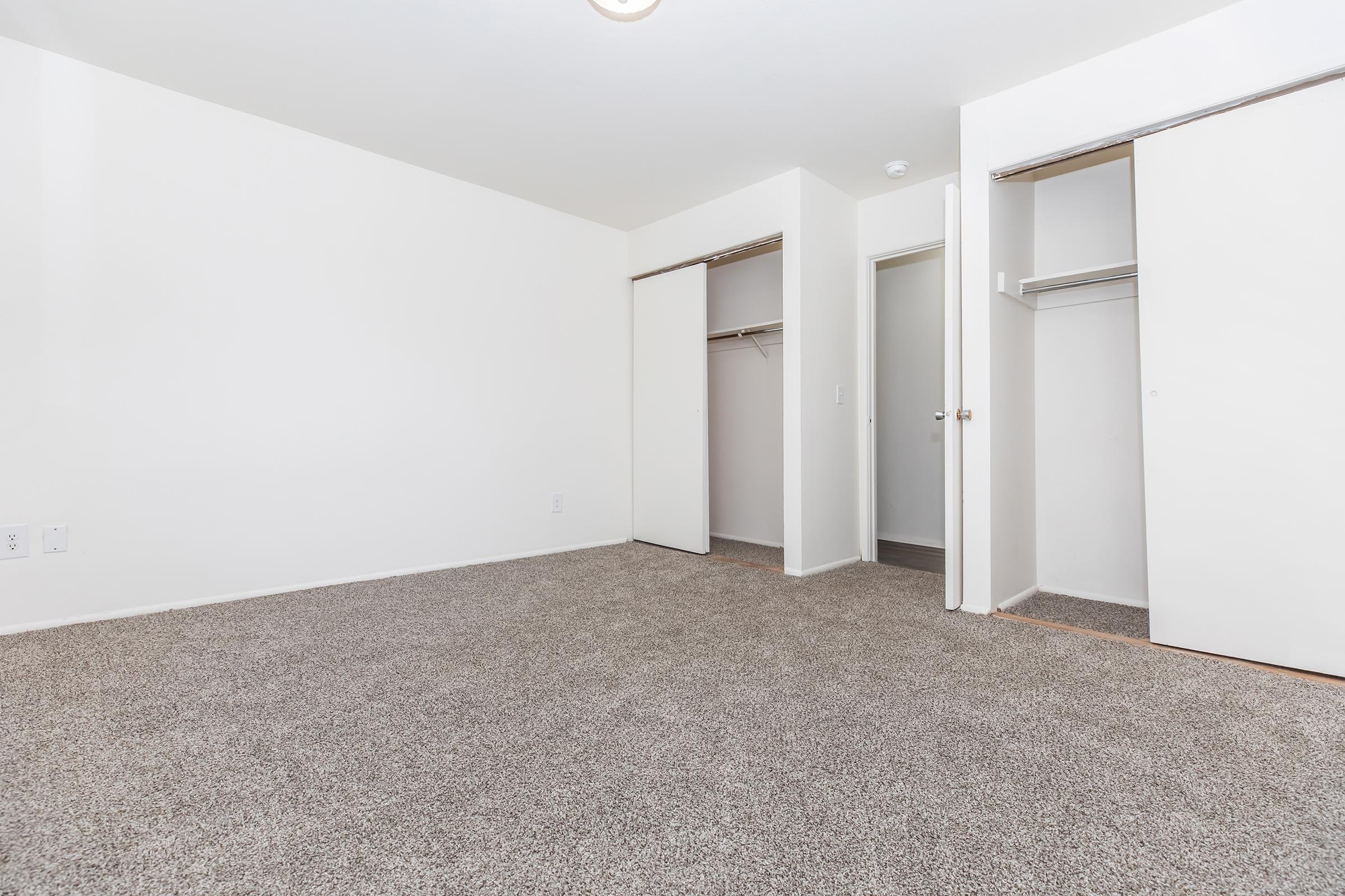 A vacant room featuring light-colored walls, beige carpet flooring, and two open closets on the left side. The layout is simple, with a door leading to another room in the background. The space appears bright and airy, suitable for various uses.