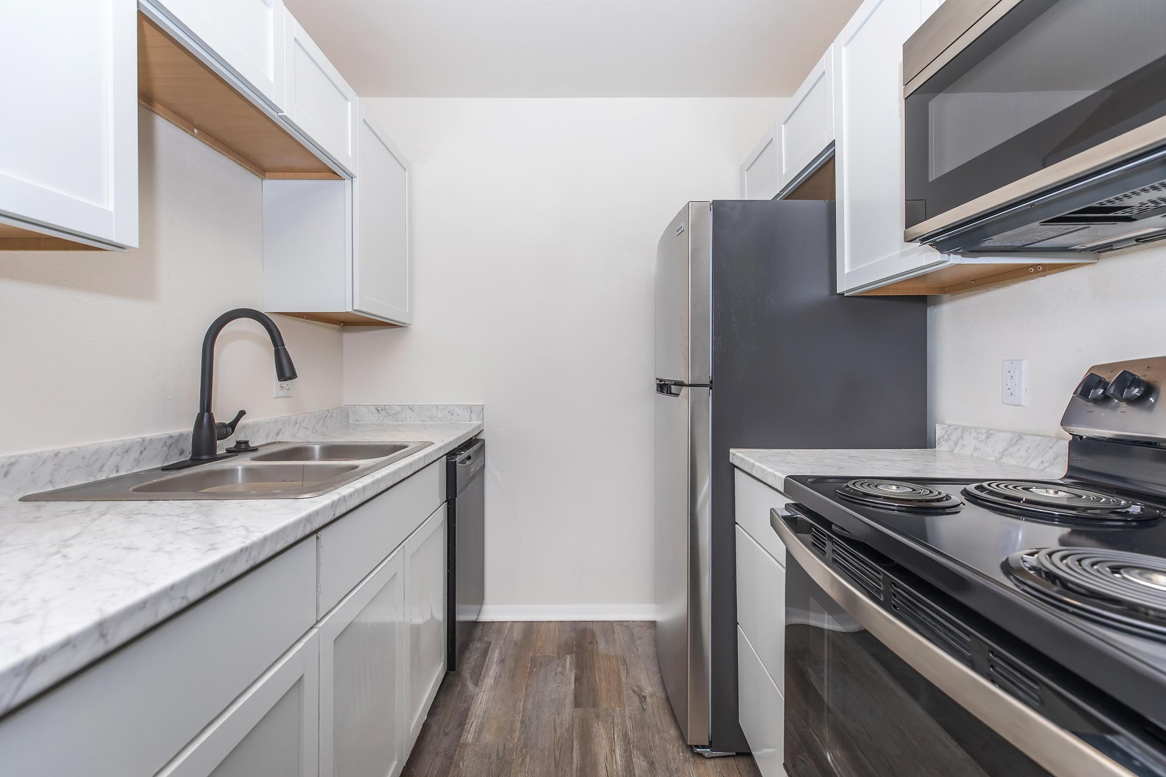 A modern kitchen featuring white cabinetry, a stainless steel refrigerator, and a black stove and microwave. The countertops are made of marble, and the flooring is wood-like. The kitchen is well-lit and organized, with a double sink and ample storage space.