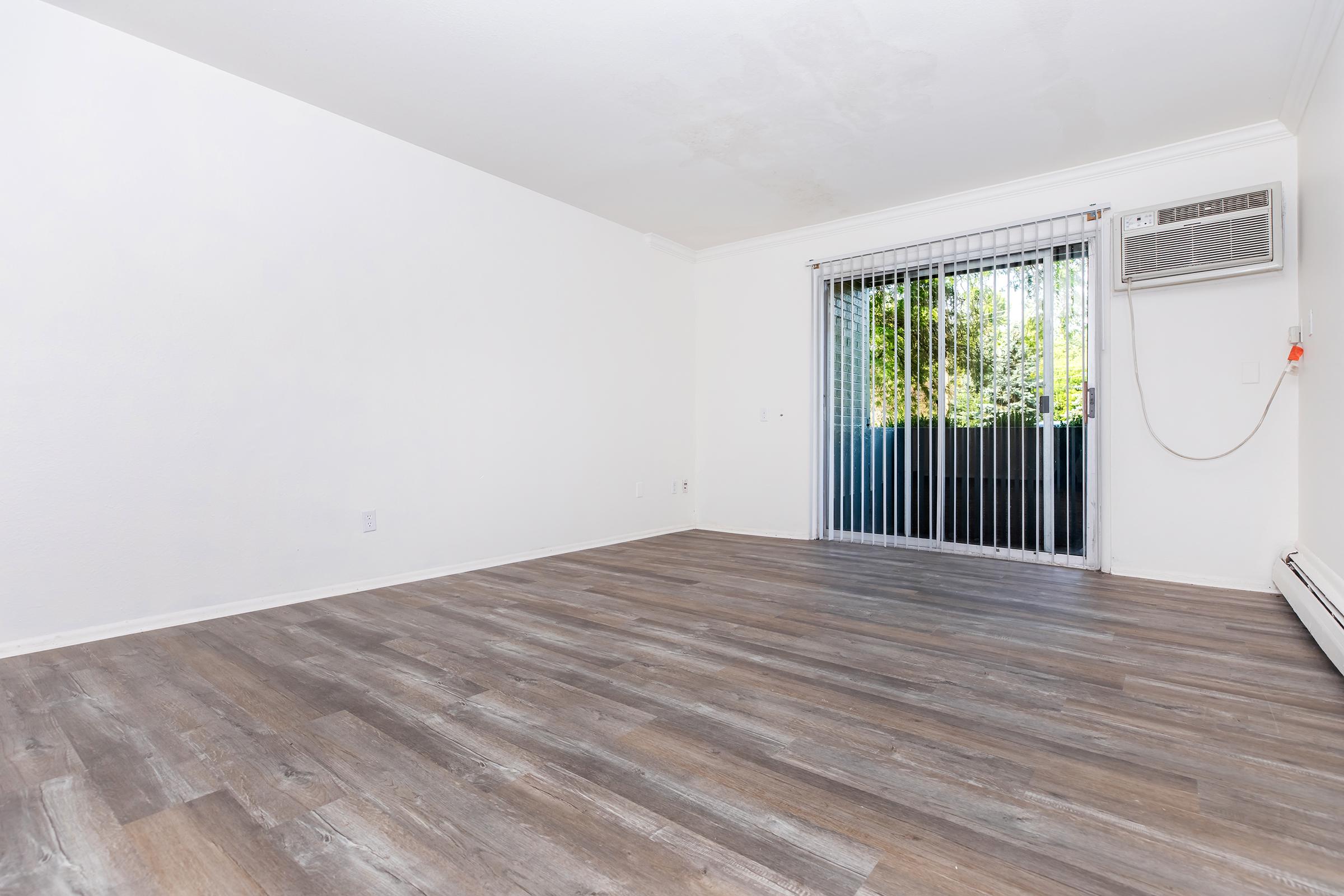 A spacious, empty room with light wood flooring and white walls. One side features a sliding glass door leading to an outdoor area, while an air conditioning unit is mounted on the wall. The overall atmosphere is clean and bright, suitable for various uses or furniture arrangements.