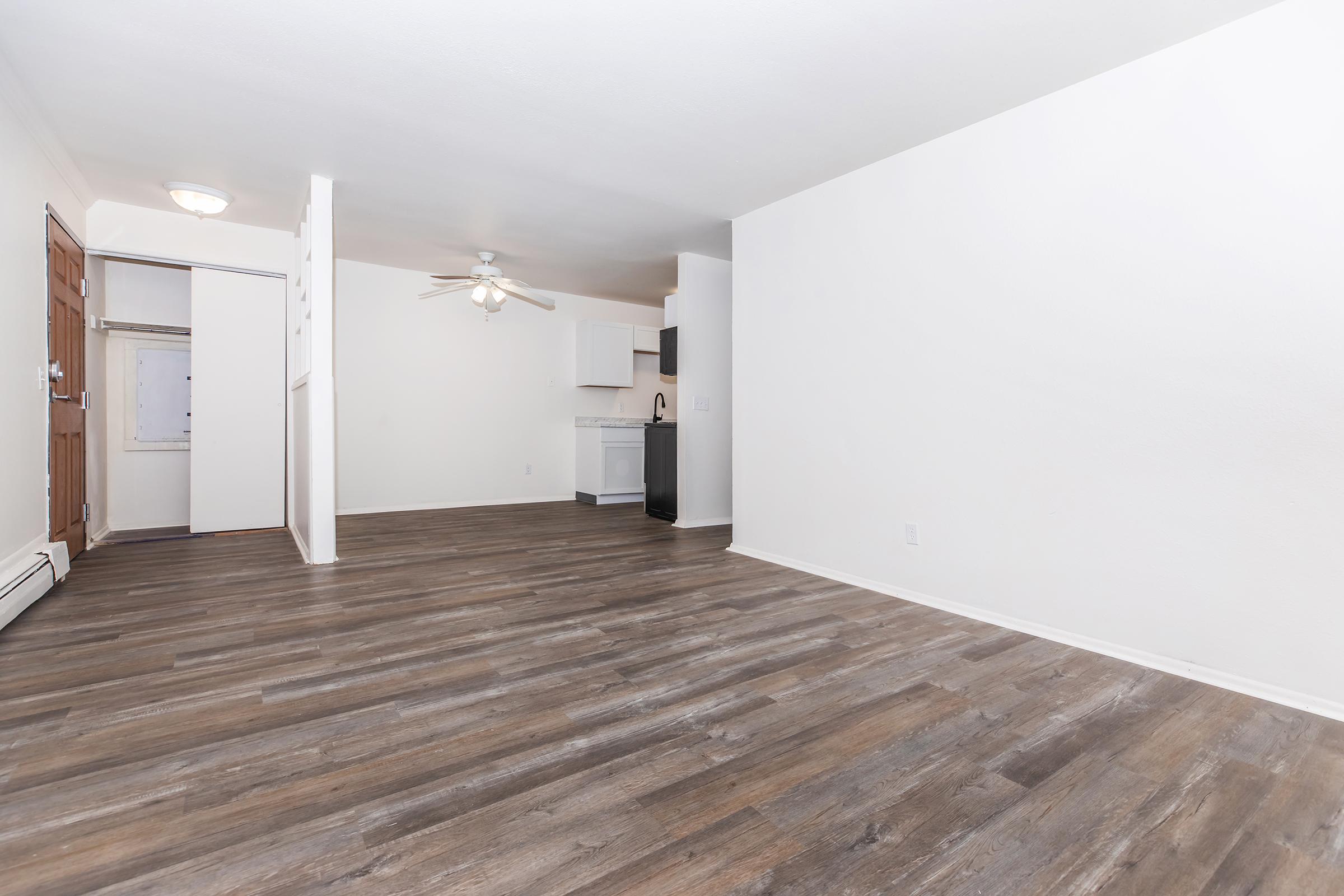 Spacious and bright living area featuring hardwood-style flooring, a ceiling fan, and an open layout. The room connects to a simple kitchen with white cabinetry and appliances, providing a cozy, inviting space ideal for relaxation or entertaining.