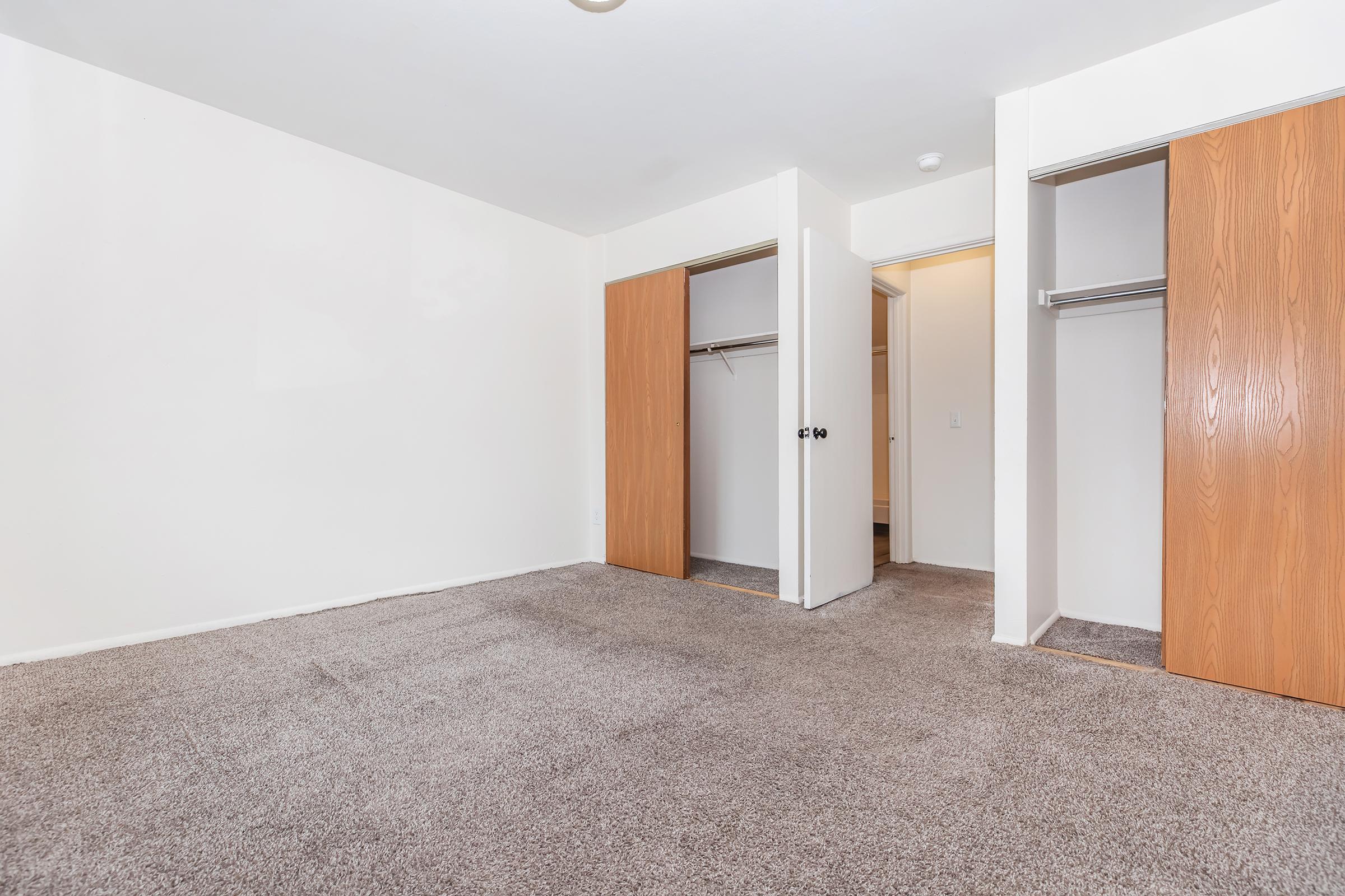 Spacious, empty bedroom featuring light-colored walls and a carpeted floor. Two closed closet doors on the left and an open door on the right leading to another room. Natural light is visible, creating a bright and airy atmosphere. Ideal for personalization and decorating.