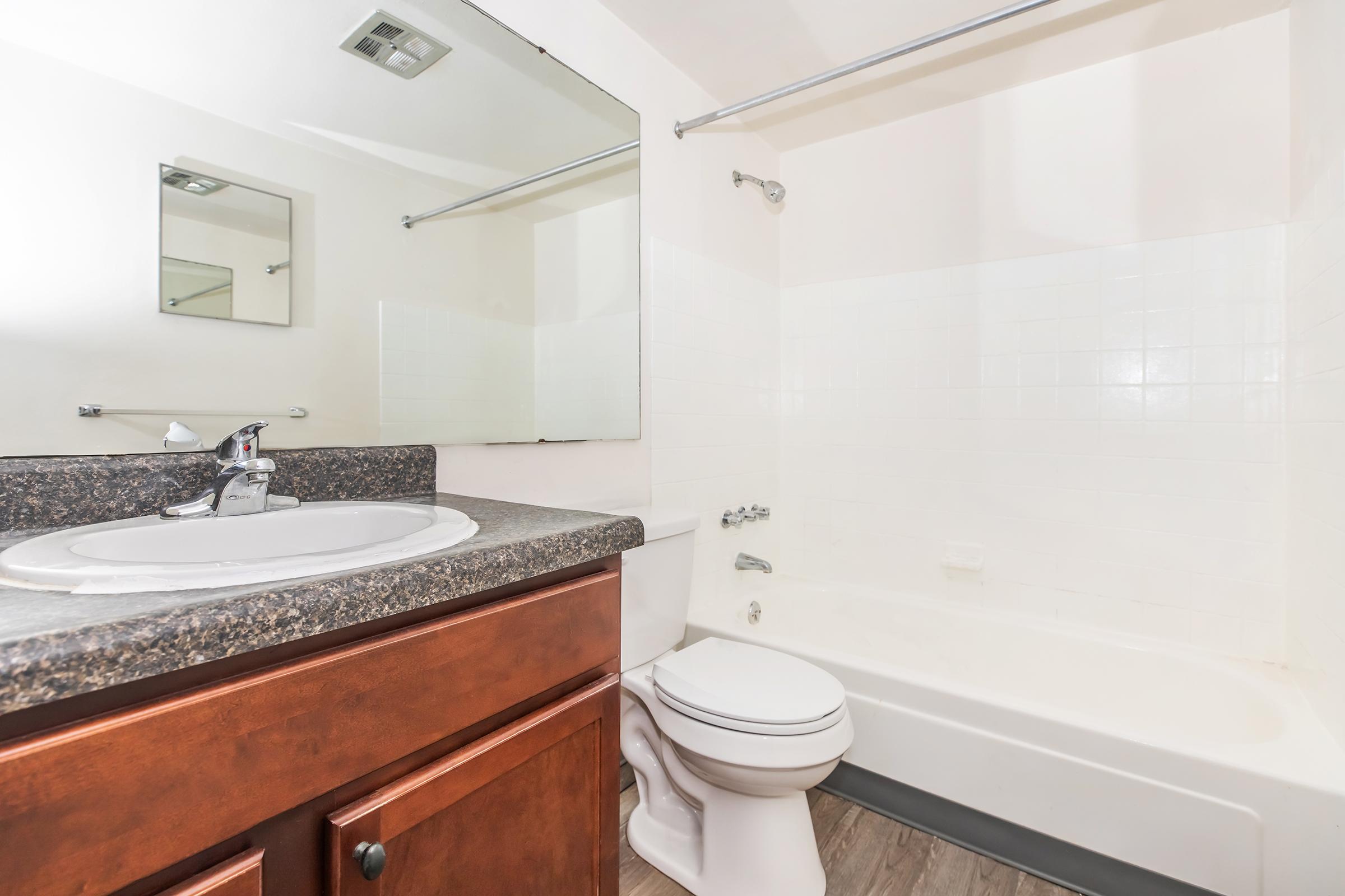 A clean bathroom featuring a white bathtub with a shower, a sink with a dark granite countertop, and a large mirror. The walls are painted light, and there is a wooden cabinet under the sink. The floor is tiled.